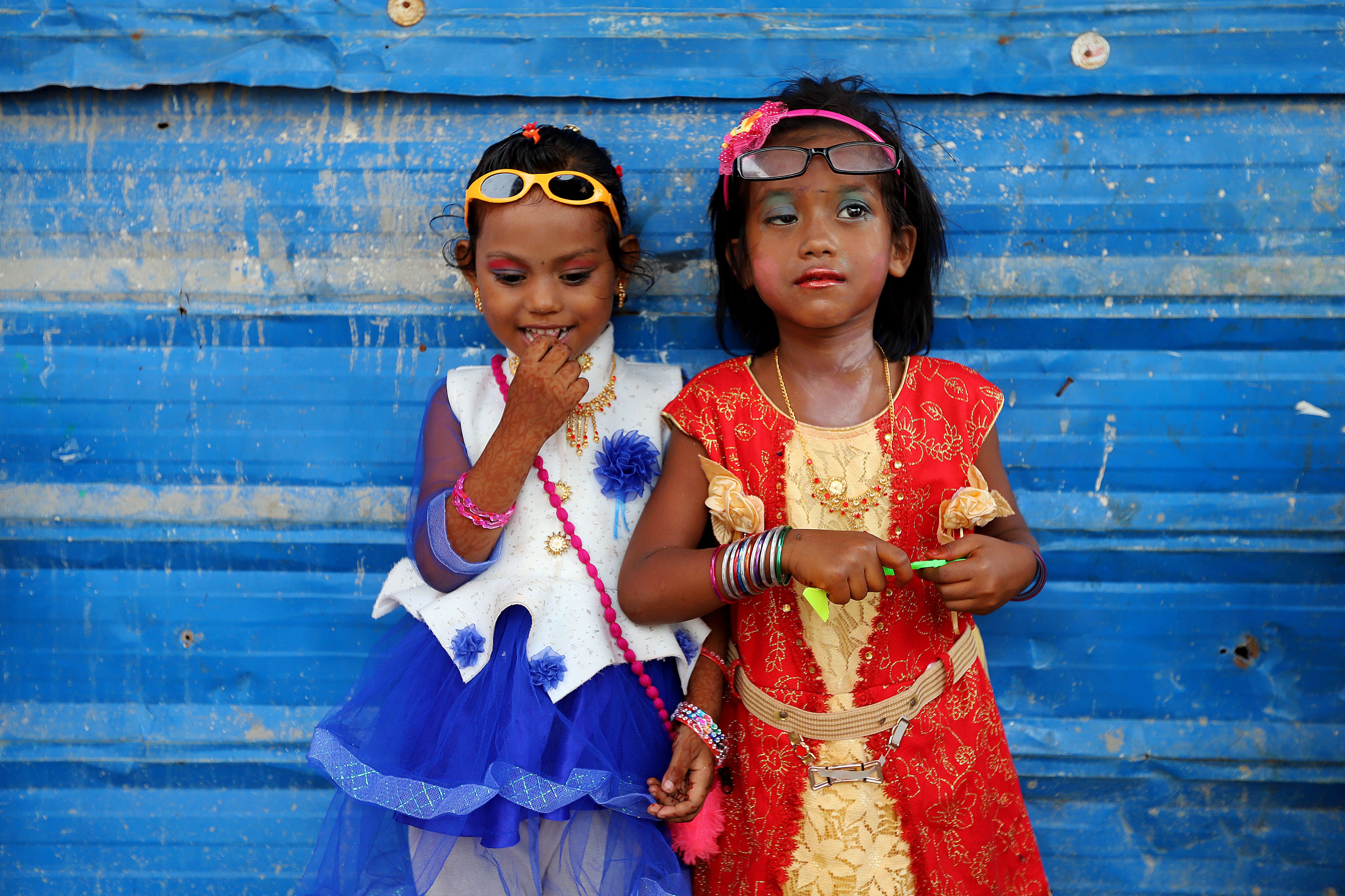 Two young girls in makeup and dresses lean against a blue wall 