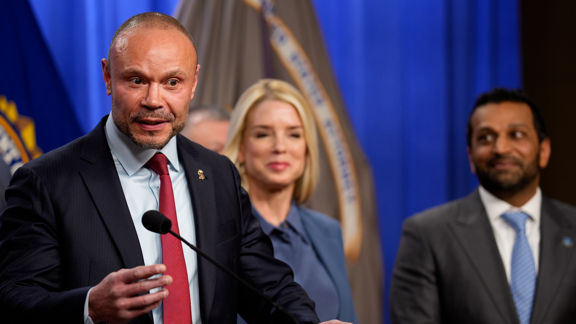 Bongino wearing a dark blue suit jacket, light blue shirt and red tie speaking at a podium