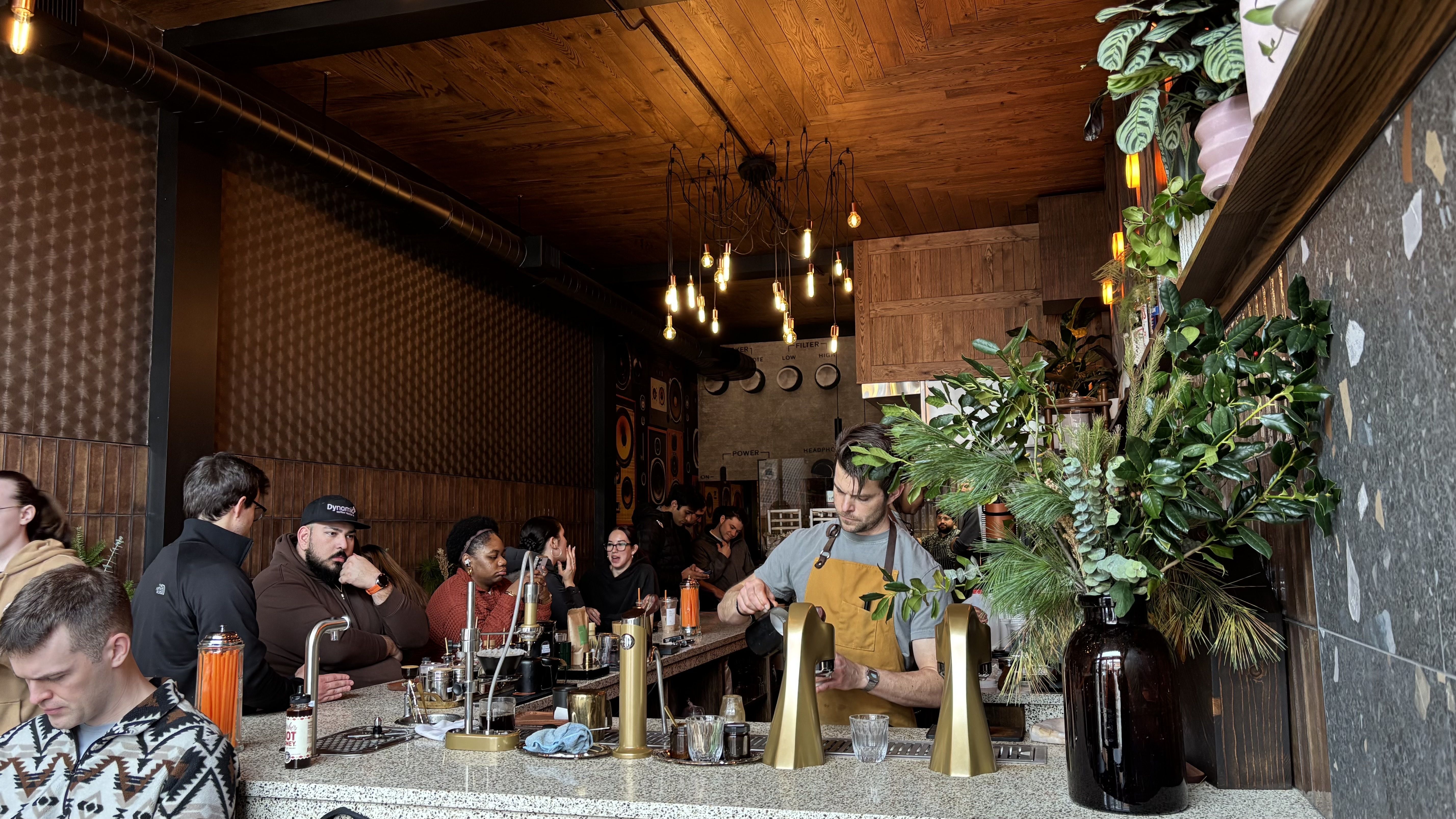 A barista in a yellow apron prepares drinks behind a counter in a cozy coffee shop with wooden ceiling, hanging lights, green plants, and customers seated at the counter.