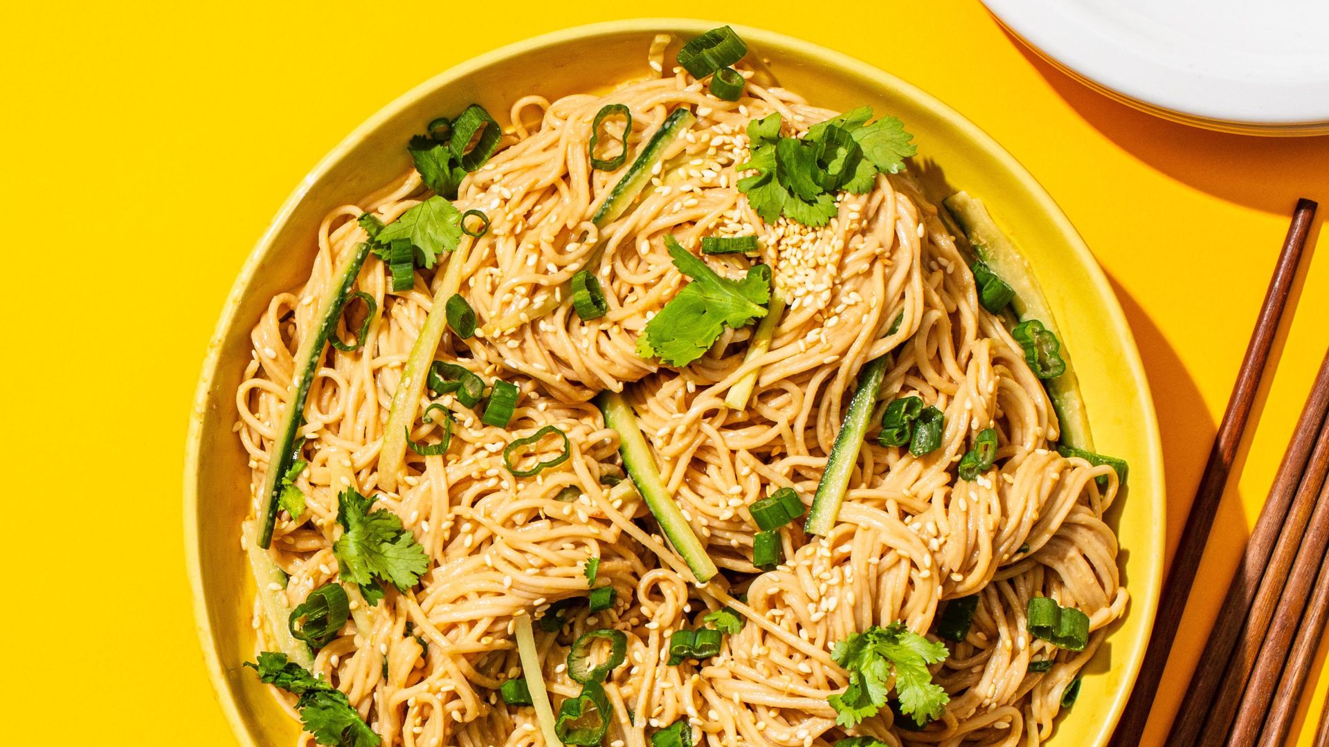 An aerial shot of a yellow bowl of sesame noodles, with cilantro leaves scattered on top, sitting on a yellow table.