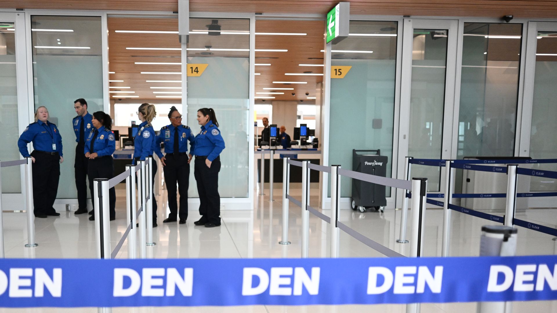 Multiple TSA agents stand near a clear door behind a blue tape with the word DEN inside an airport.