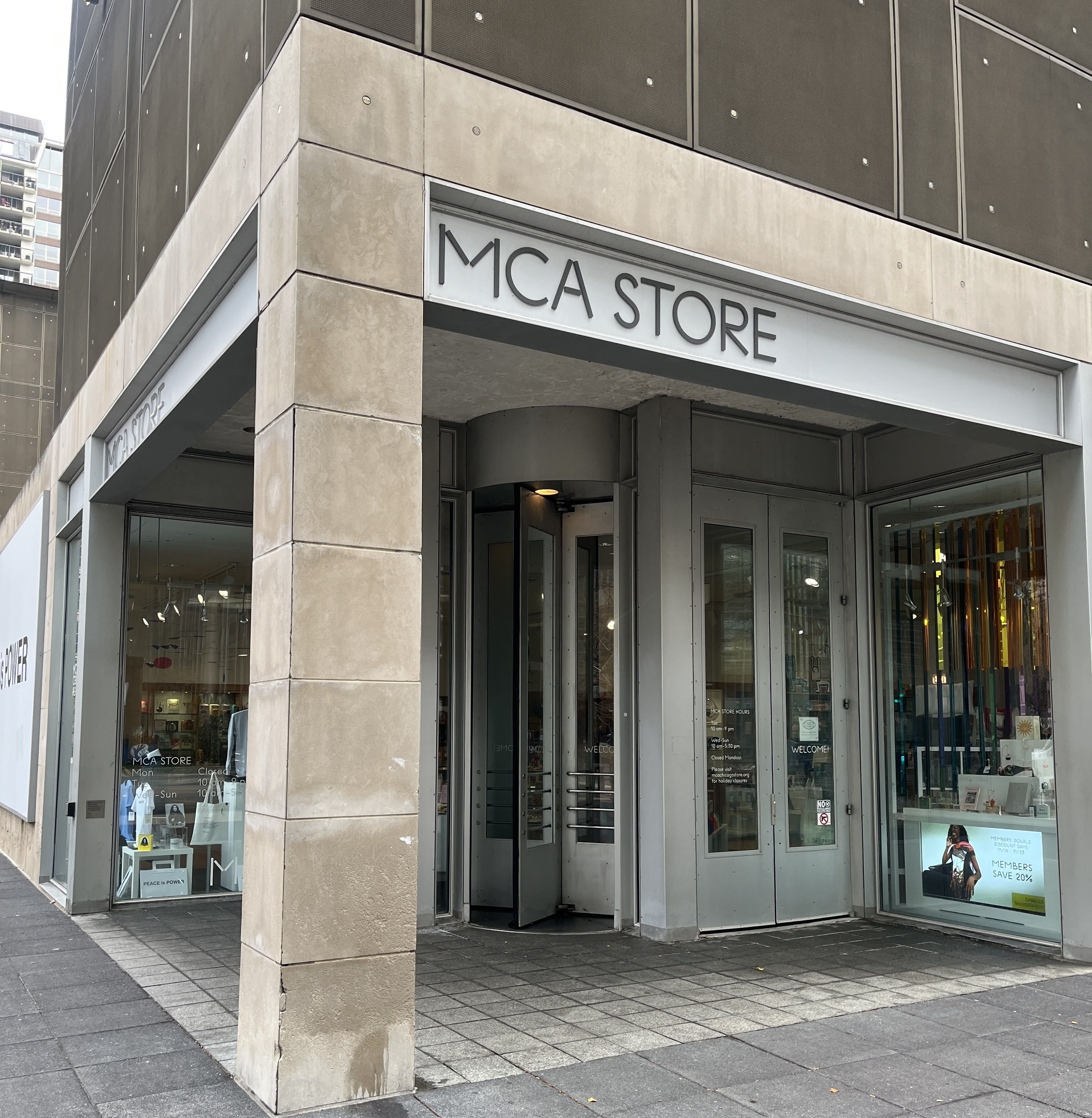 Exterior view of MCA Store with beige stone pillars, revolving entrance door, glass windows showing displays and a digital sign offering 20% member discount.