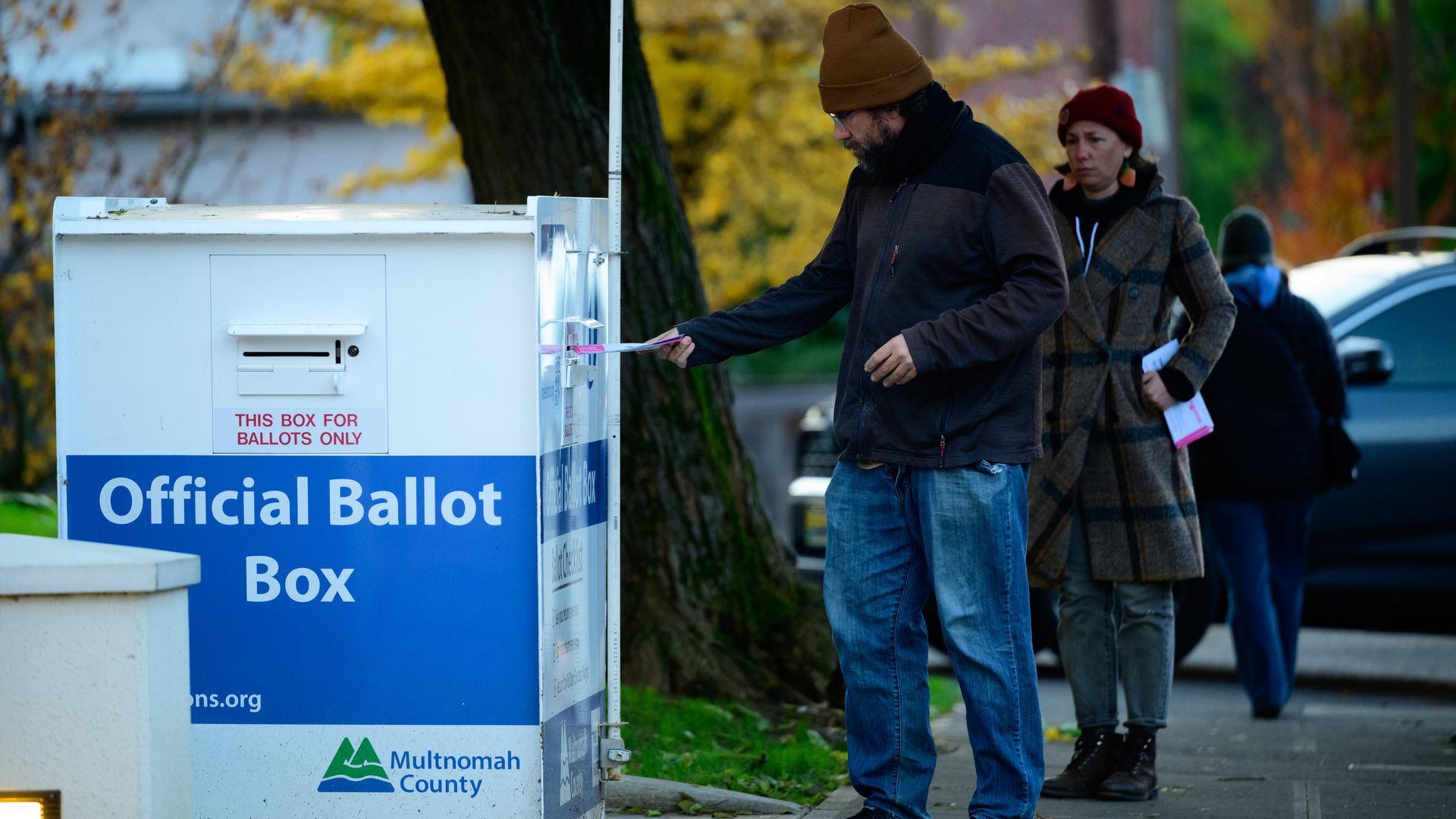 Man in a brown beanie inserts a ballot into a white official ballot box; a woman in a red hat stands behind them as autumn leaves and trees fill the background.