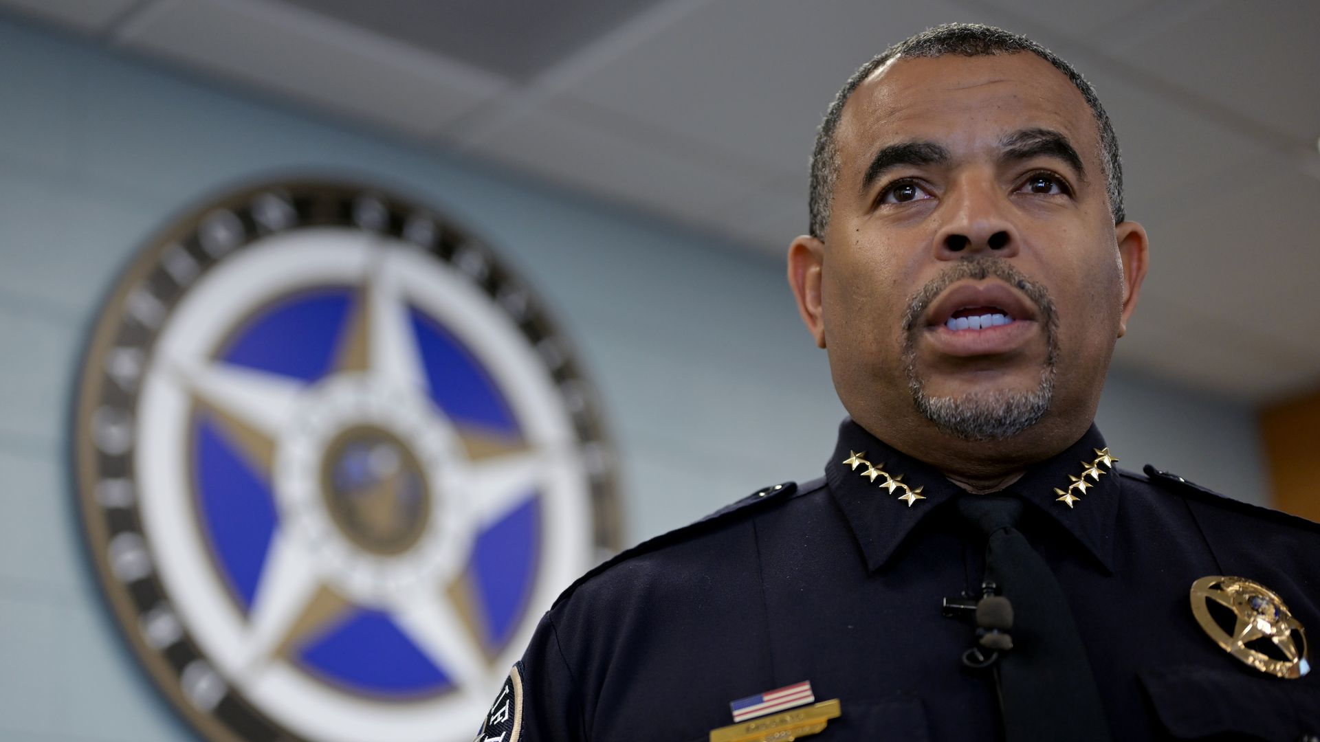 A uniformed law enforcement officer in a black uniform speaks in the foreground, while a large, blue and white badge is blurry in the background. 
