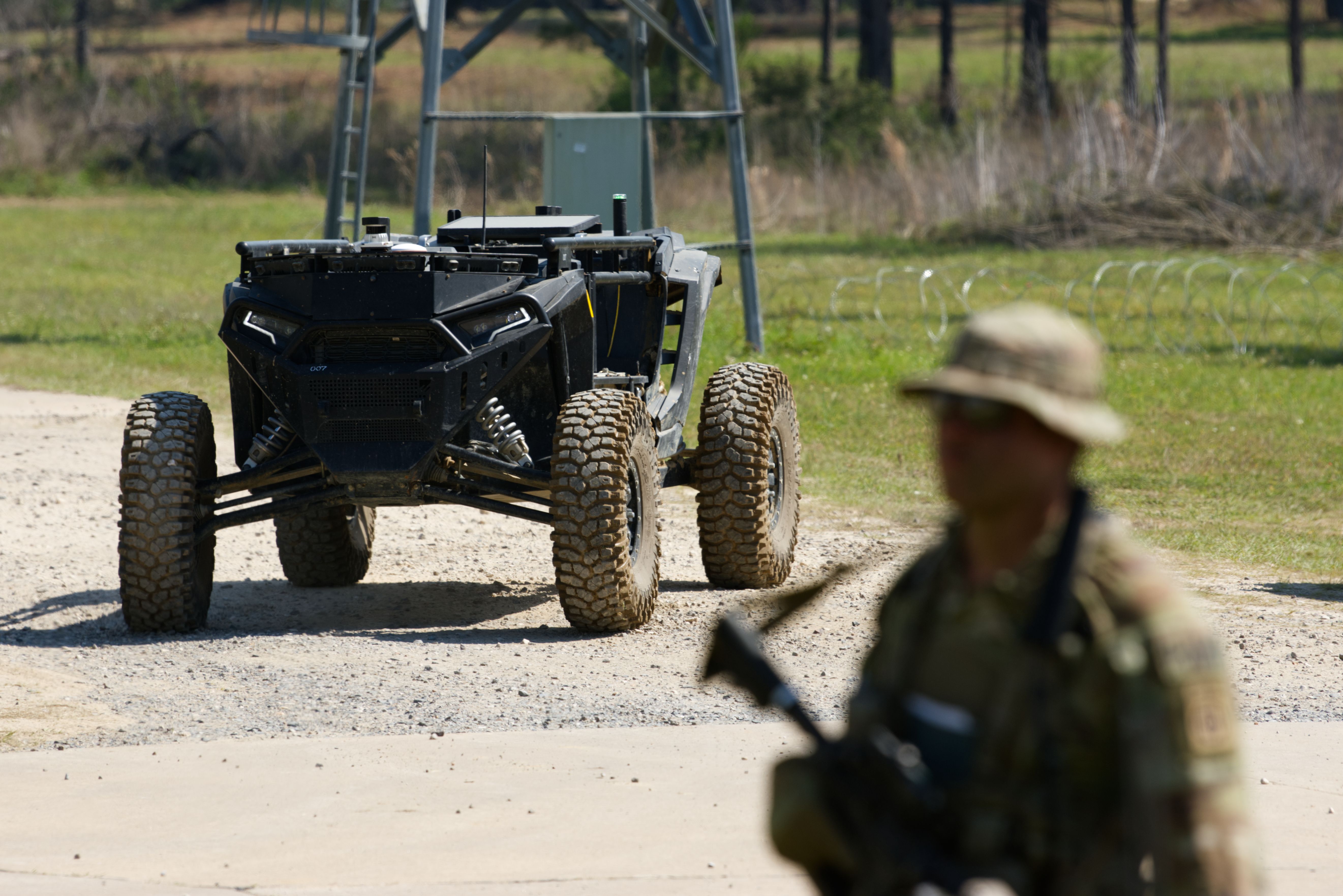 Black off-road UTV with oversized tires sits on a dirt road. A blurred camouflaged soldier stands in the foreground.