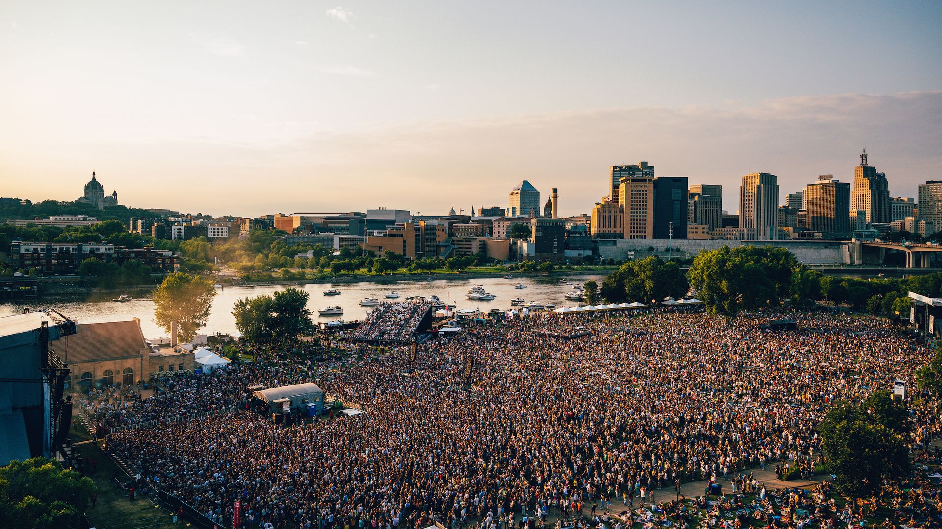 Wide city skyline at sunset with a large crowd gathered at an outdoor venue along the river. Modern high-rises to the right, a domed historic building on the left, boats on the water, warm golden sky.