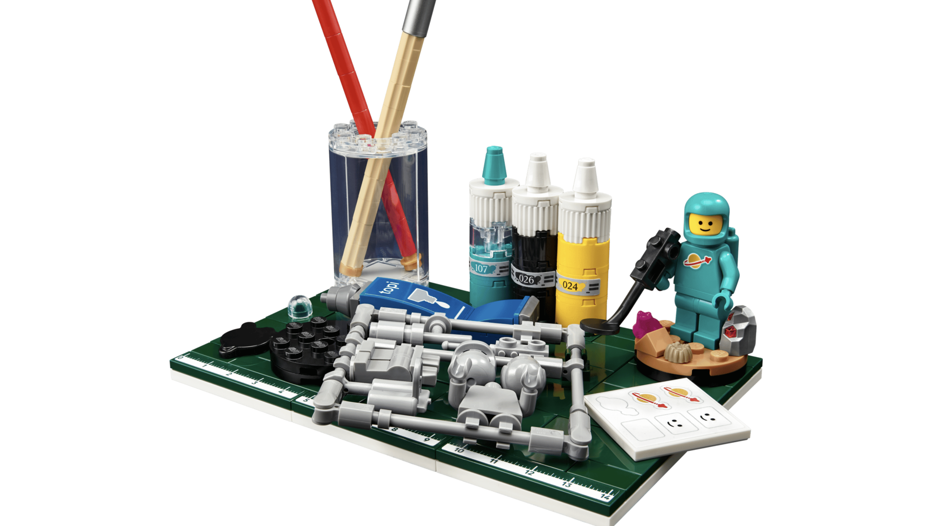 LEGO workstation with two brushes in a clear holder, teal-capped paint bottles, a green baseplate with gray parts, a teal space-suited minifigure, and a ruler.