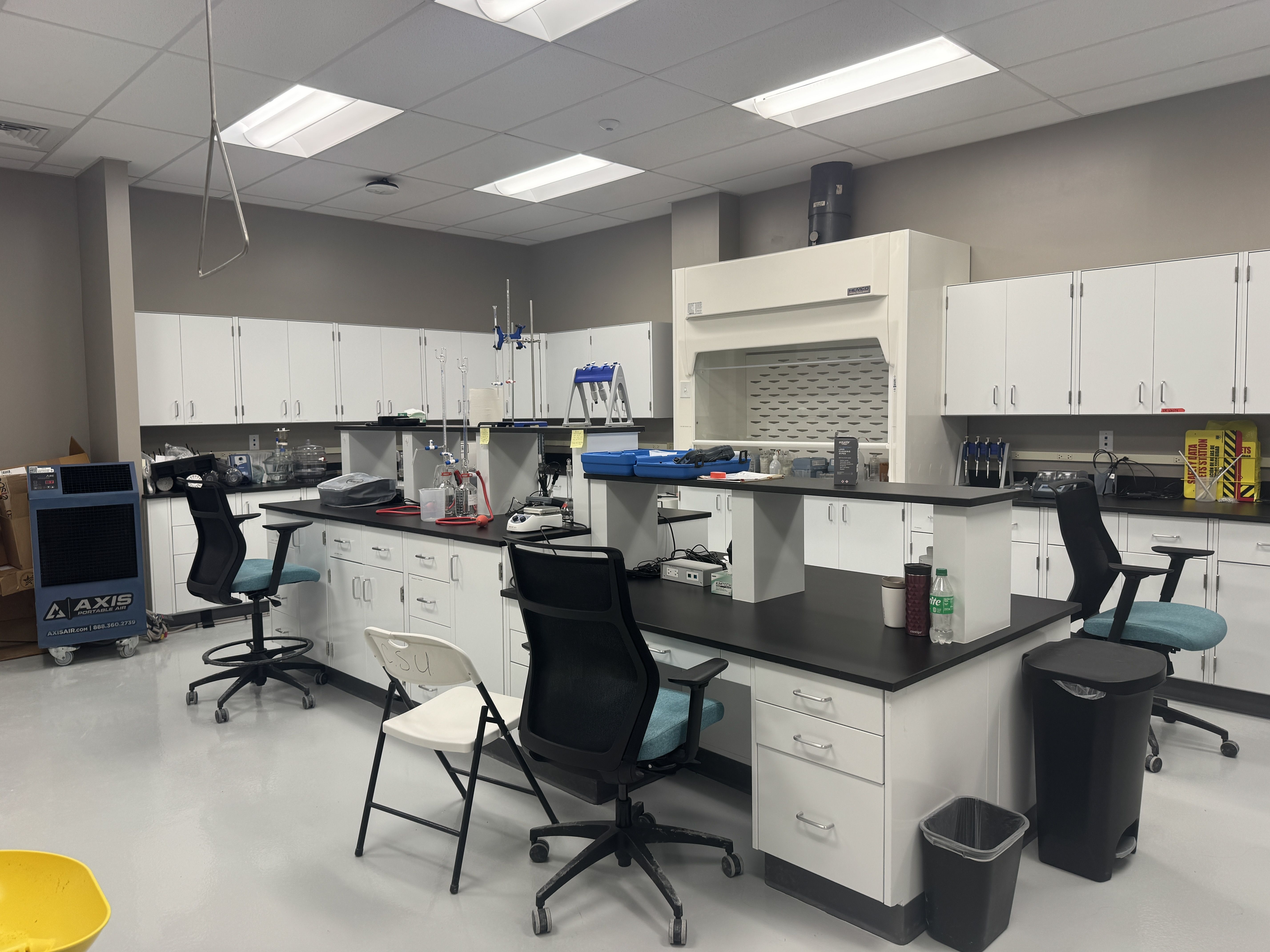 Modern laboratory with white cabinets, black countertops, and multiple workstations. Lab equipment, stools, and a fume hood beneath bright ceiling lights.