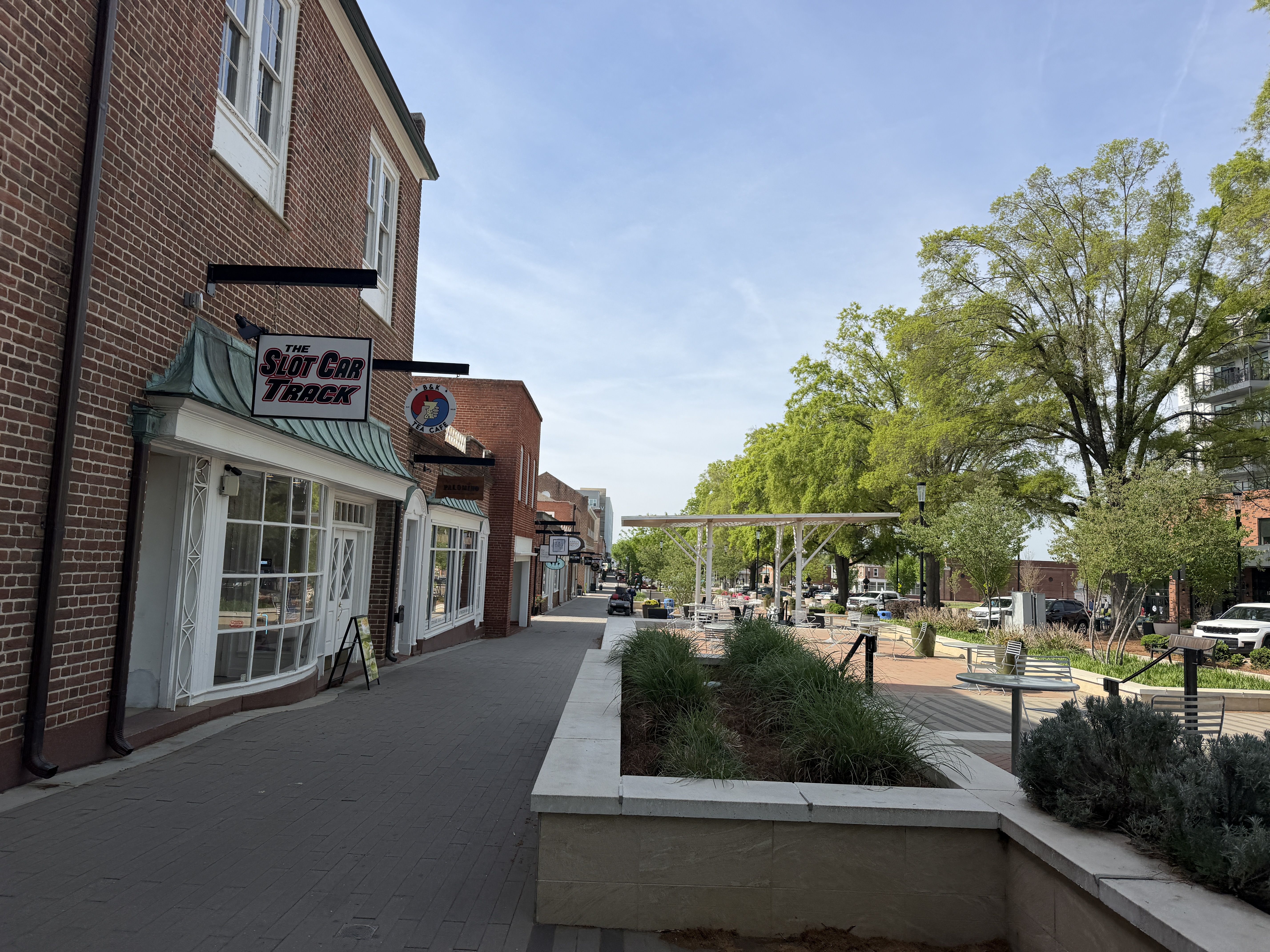 Sunny street with red brick storefronts along a brick sidewalk. A sign says "The Slot Car Track" above a shop. On the right, a landscaped plaza with trees, benches, and a white pergola.