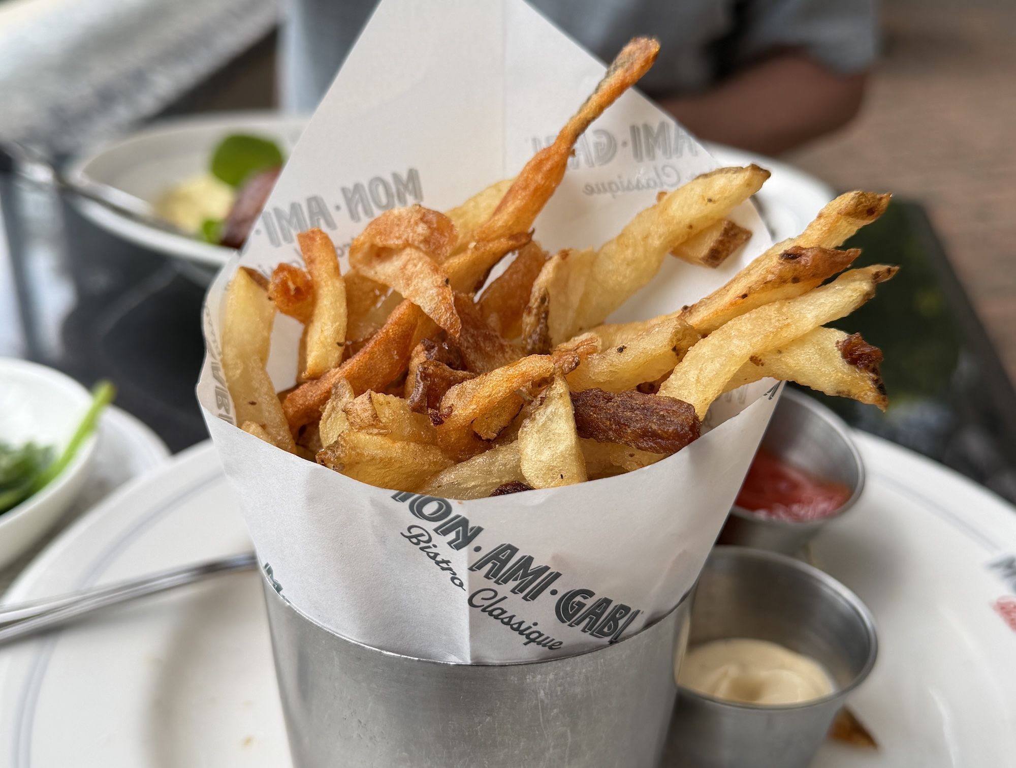 Crispy French fries spill from a white paper cone placed in a metal cup; a red dipping sauce cup and a blurred plate with greens are in the background.