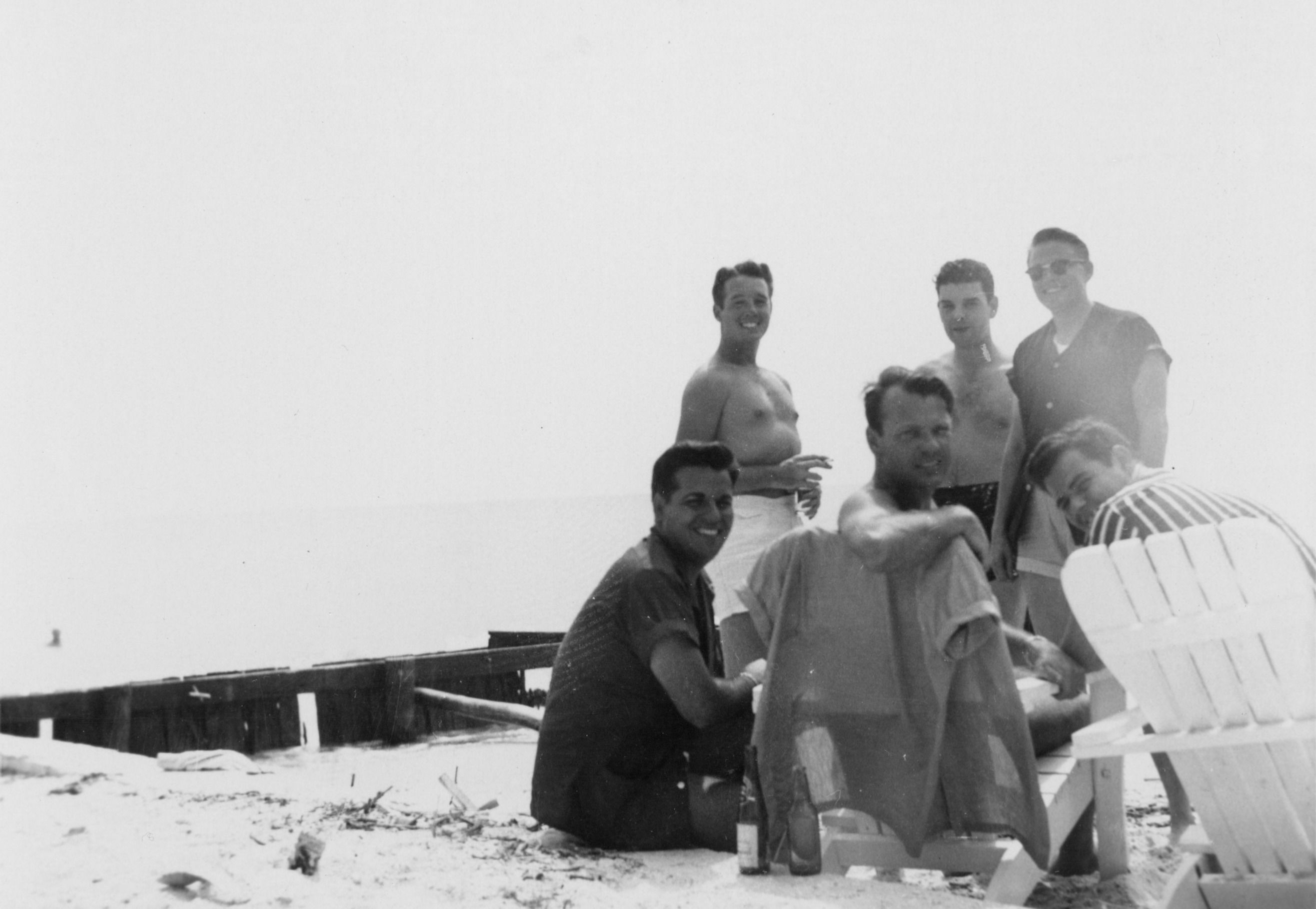 a black and white photo of a group of people, some with shirts off and some not, smiling and looking at the camera. they're at the beach