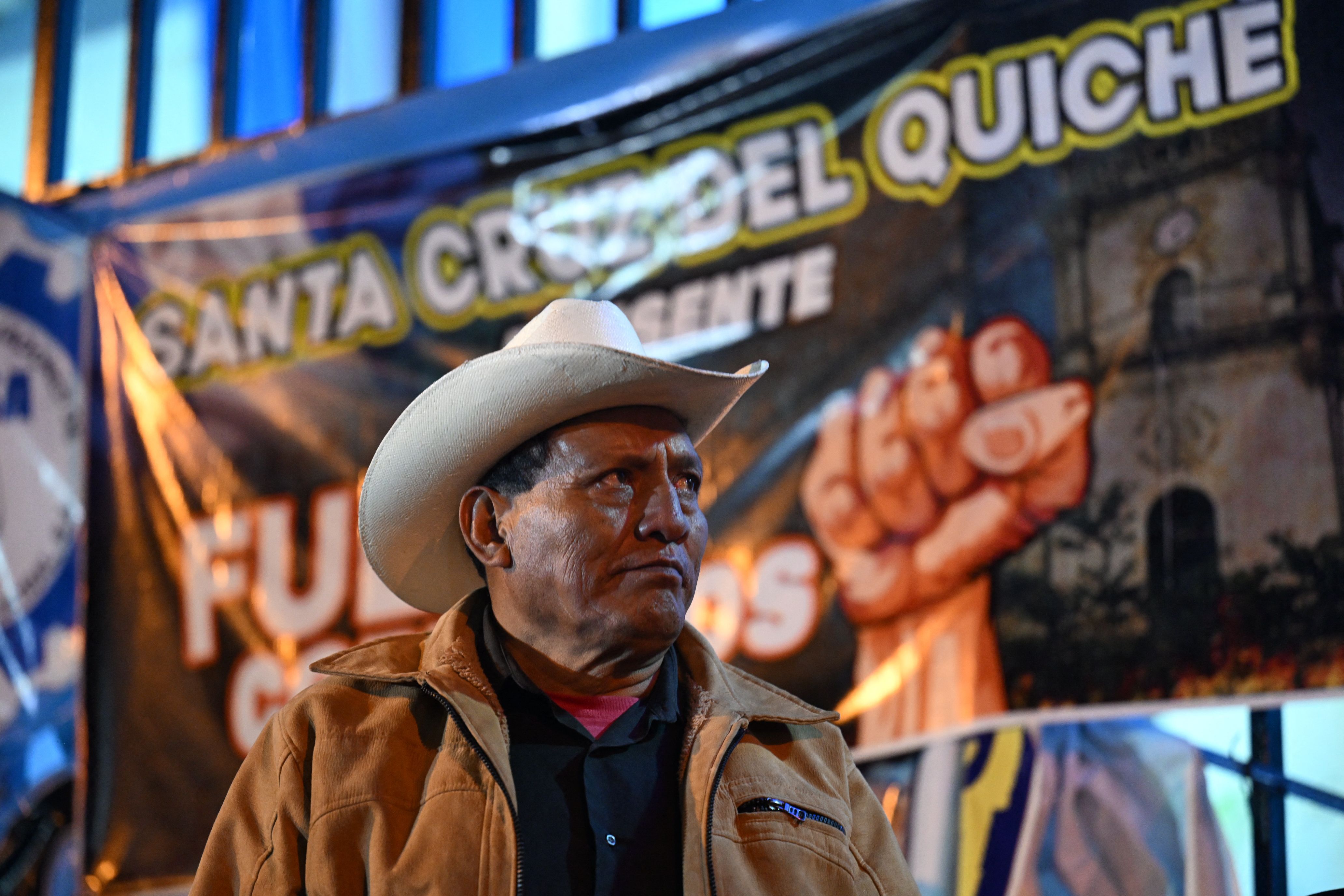a man in a cowboy hat looks off to the side during a protest against the Guatemalan attorney general. He is wearing a brown jacket and stands in front of a large banner with a picture of a fist 
