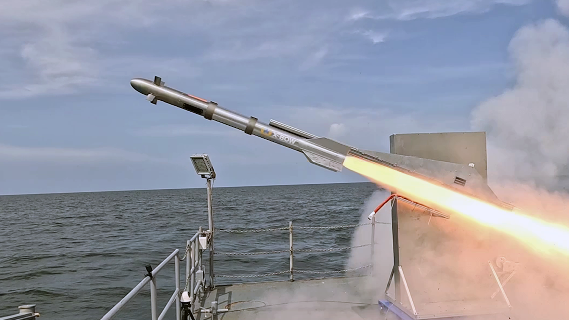 A rocket launch from a seaside test platform. A gray cylindrical missile with fins angles upward. Orange flames and smoke shoot out at the base.