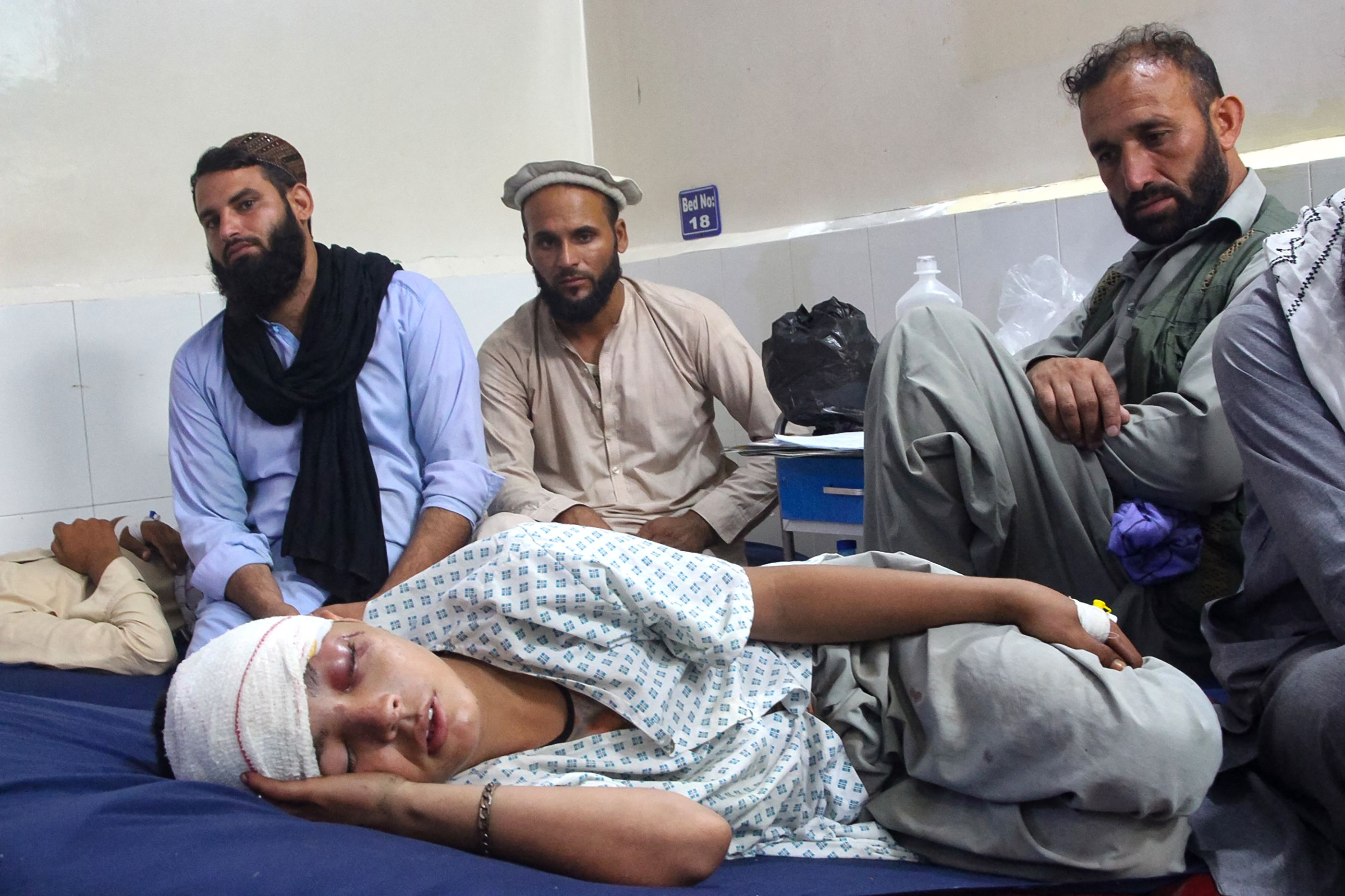 An injured Afghan boy with a bandage on his head and left hand receiving treatment at a hospital as three men in traditional Afghan dress look on.