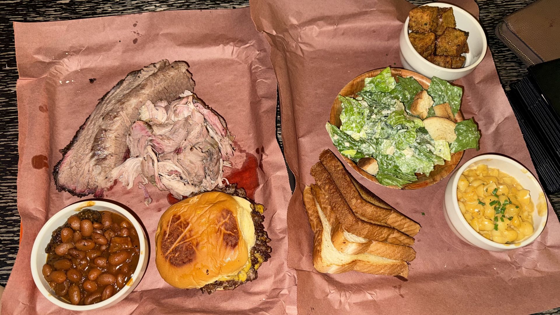Barbecue plate with smoked brisket, pulled pork, cheeseburger, baked beans, Caesar salad, toasted bread slices, crispy potato cubes, and creamy macaroni and cheese on brown paper.