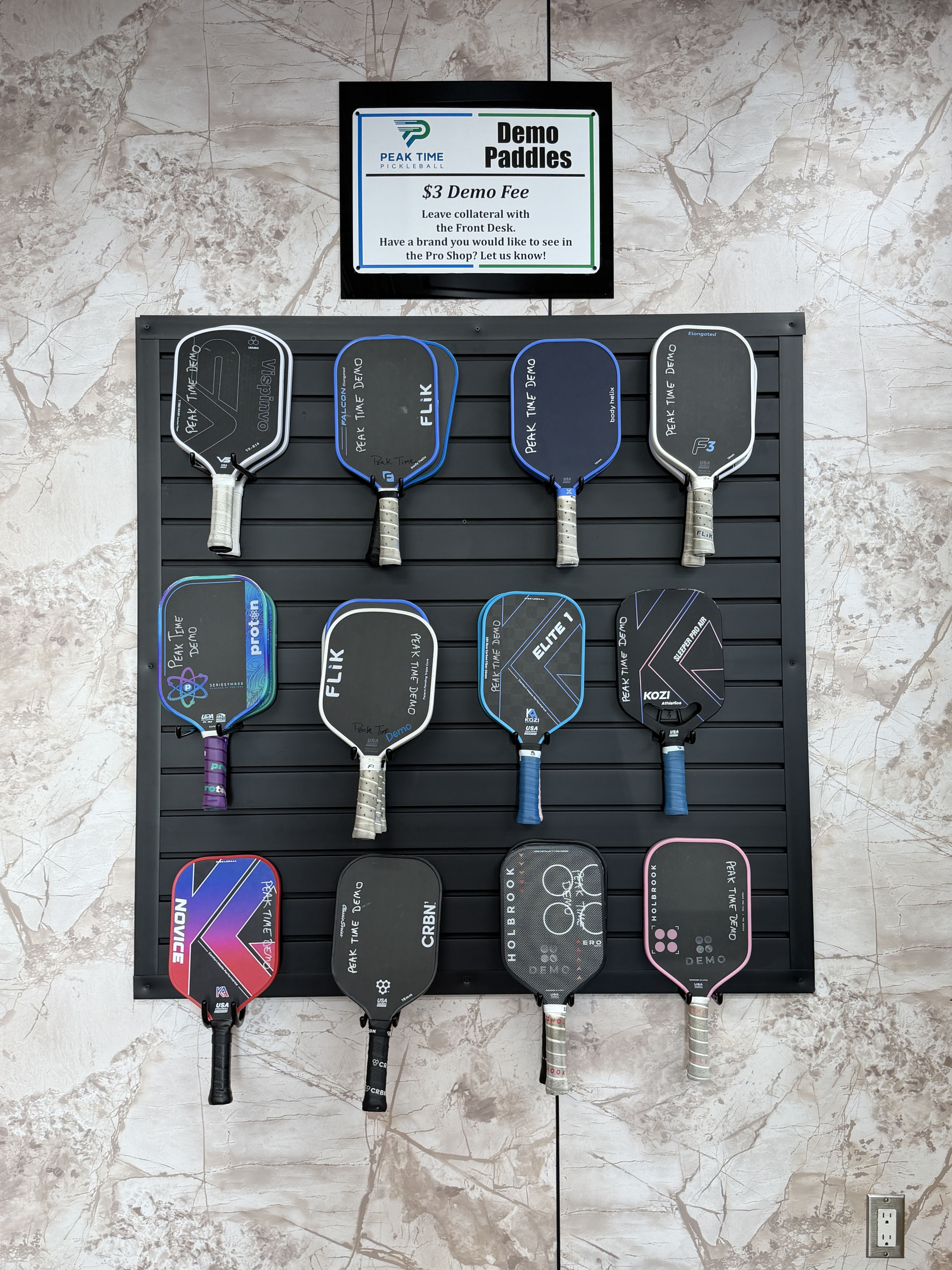 Row of pickleball paddles hanging on a dark slat wall beneath a sign reading Demo Paddles with a $3 demo fee; paddles in blue, gray, black, and purple against a marble backdrop.