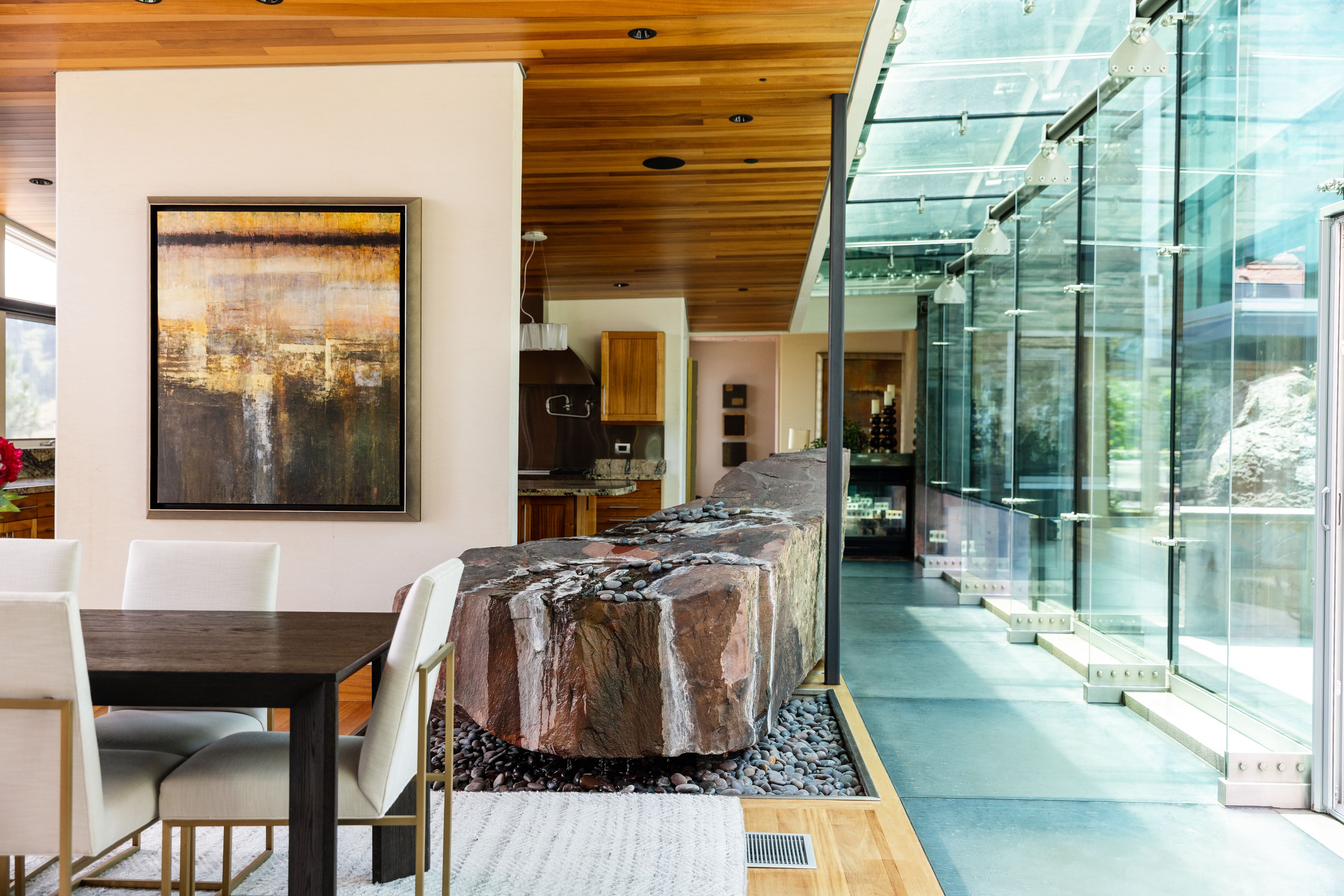 Modern dining area with white chairs and dark wooden table beside a large rough stone feature surrounded by pebbles, with wood ceiling and glass wall letting in natural light.