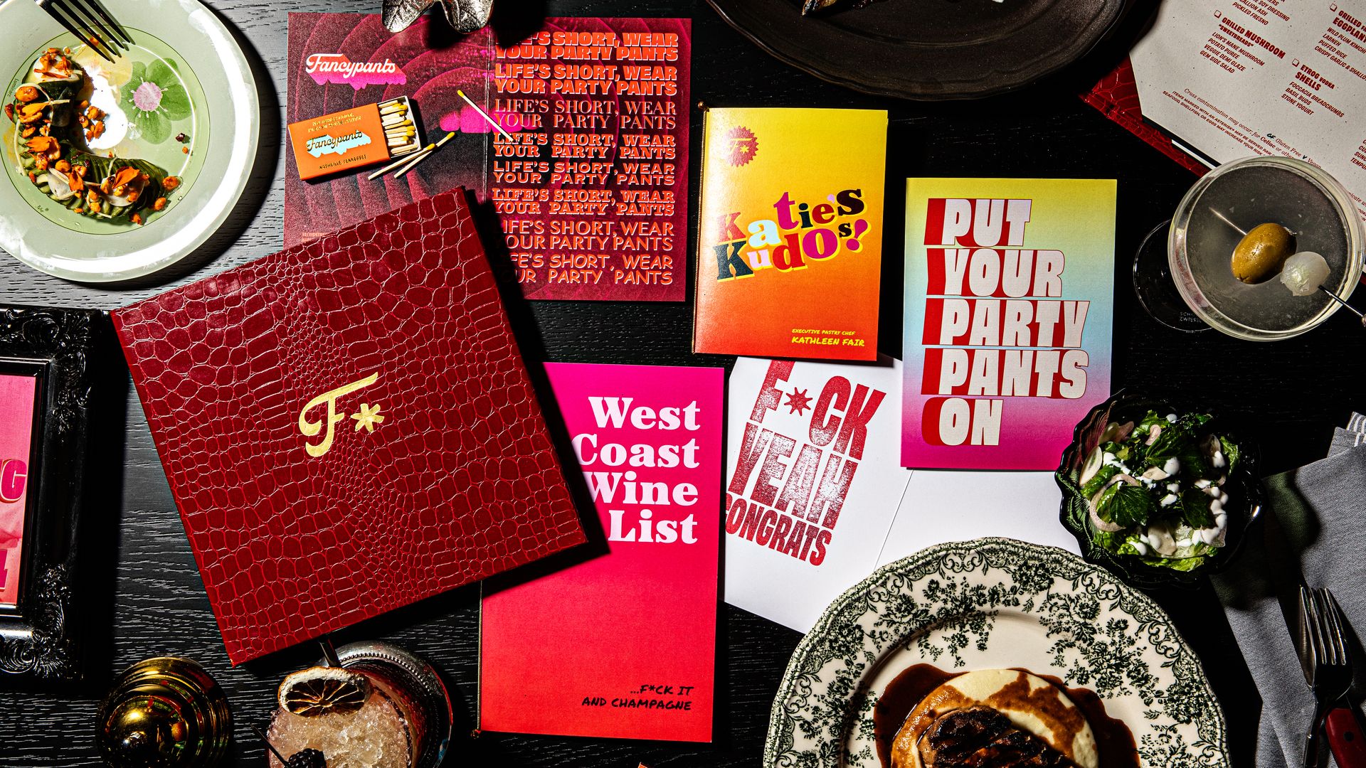 A dark table with colorful menus, including 'Katie's Kudos!' and 'Put Your Party Pants On,' dishes with gourmet food, a cocktail with olives, and a red leather menu cover with a gold F.