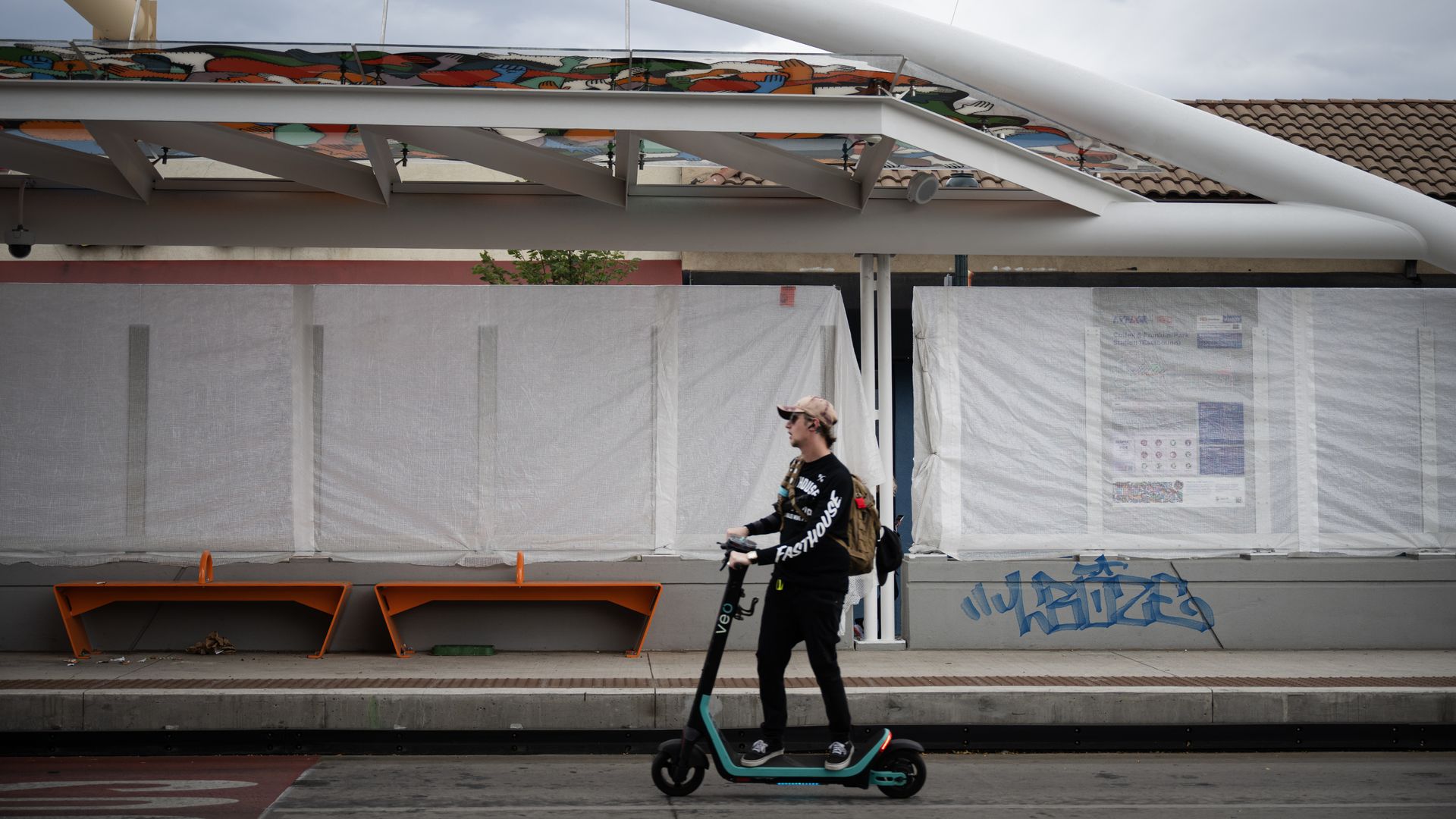 Man on a teal electric scooter rides along a street in front of a construction site with large white tubular beams and a colorful mural on an elevated platform, under a gray cloudy sky.