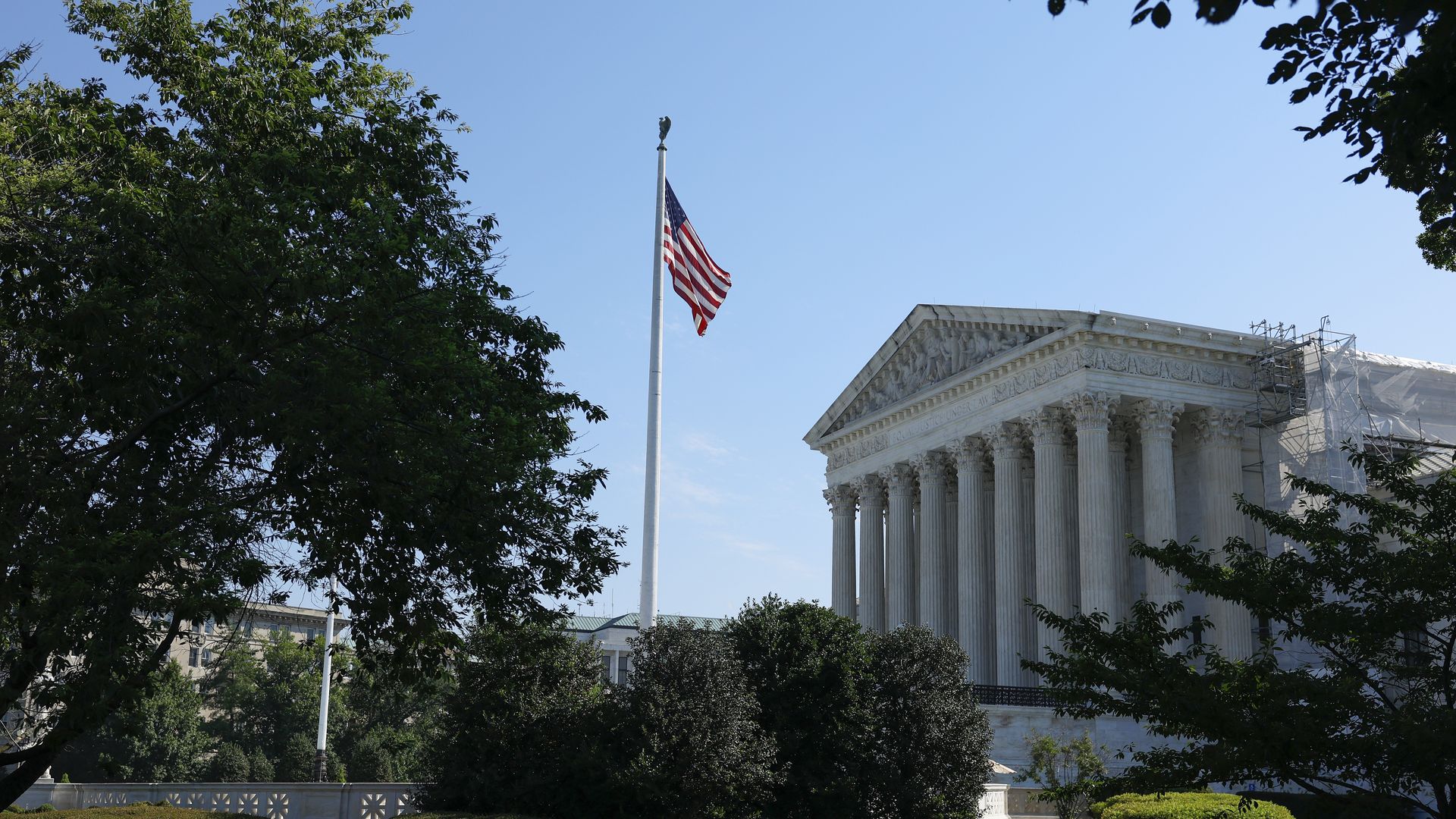 A photo of the Supreme Court on a sunny day with a blue sky. Trees are in front of it as well as an American flag in the middle. The court is to the right of the photo with trees on the left.