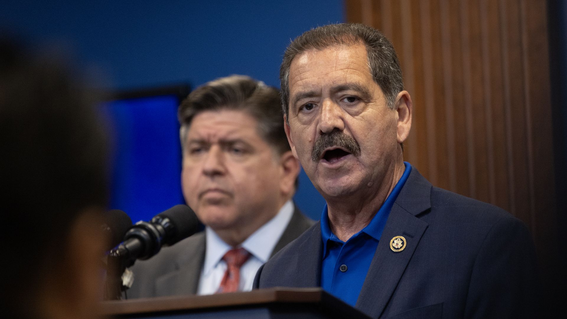 Rep. Chuy García, wearing a gray blazer over a blue polo shirt while speaking at a lectern in front of a wooden wall.