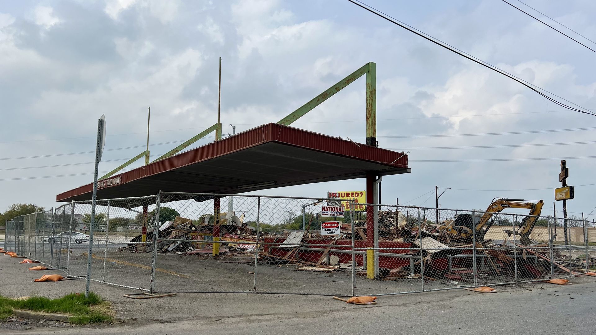 The former site of Oscar’s Taco House is seen reduced to rubble sitting behind a fence. Only a tall carport remains. A bulldozer is seen in the background under a cloudy sky.