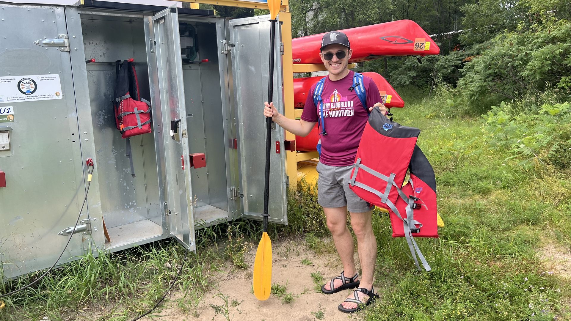 A dweedish looking man wearing an impish grin, a pinkish-purplish shirt, sunglasses, Chaco sandals, a hat with the logo of the greatest state in the world (Minnesota) on it, while holding a kayak paddle and life jacket near an aluminum locker.