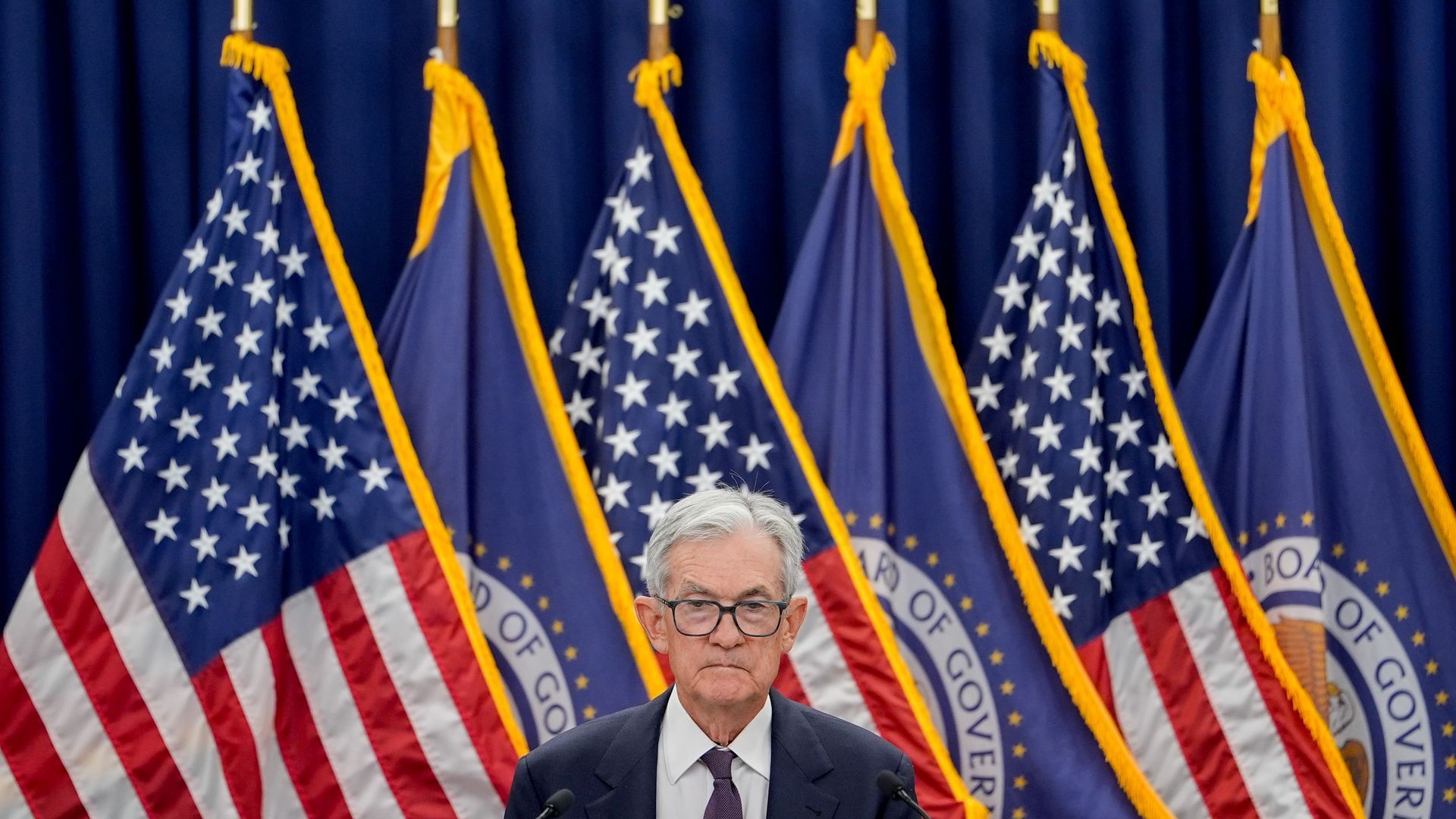 A man with gray hair and glasses in a suit stands at a podium, with several United States flags and blue government flags with gold fringe and seals behind him.