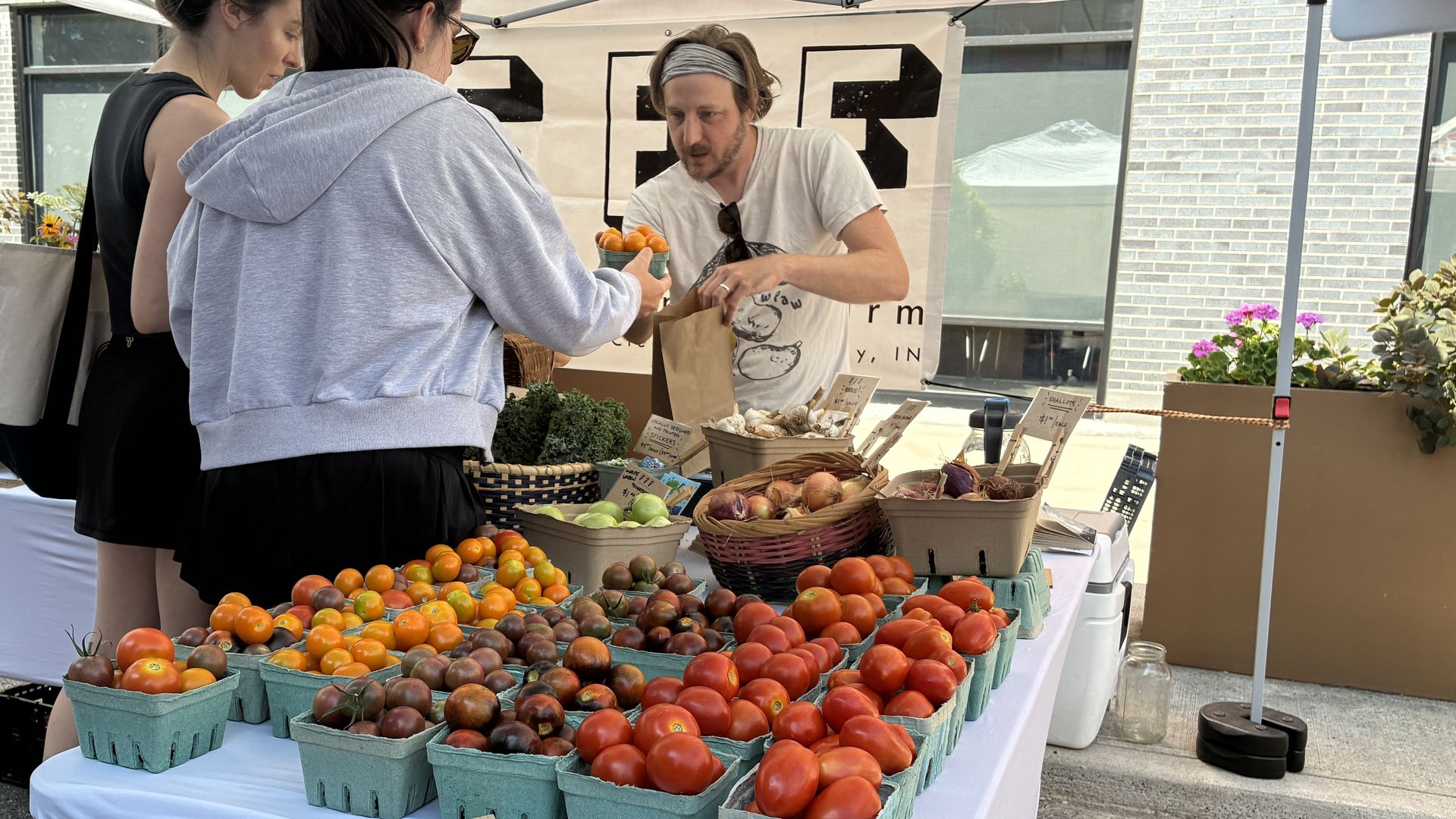 Vendor under a white canopy at an outdoor market hands a bag of tomatoes to two customers; baskets of red and orange tomatoes and handwritten price signs fill the table.