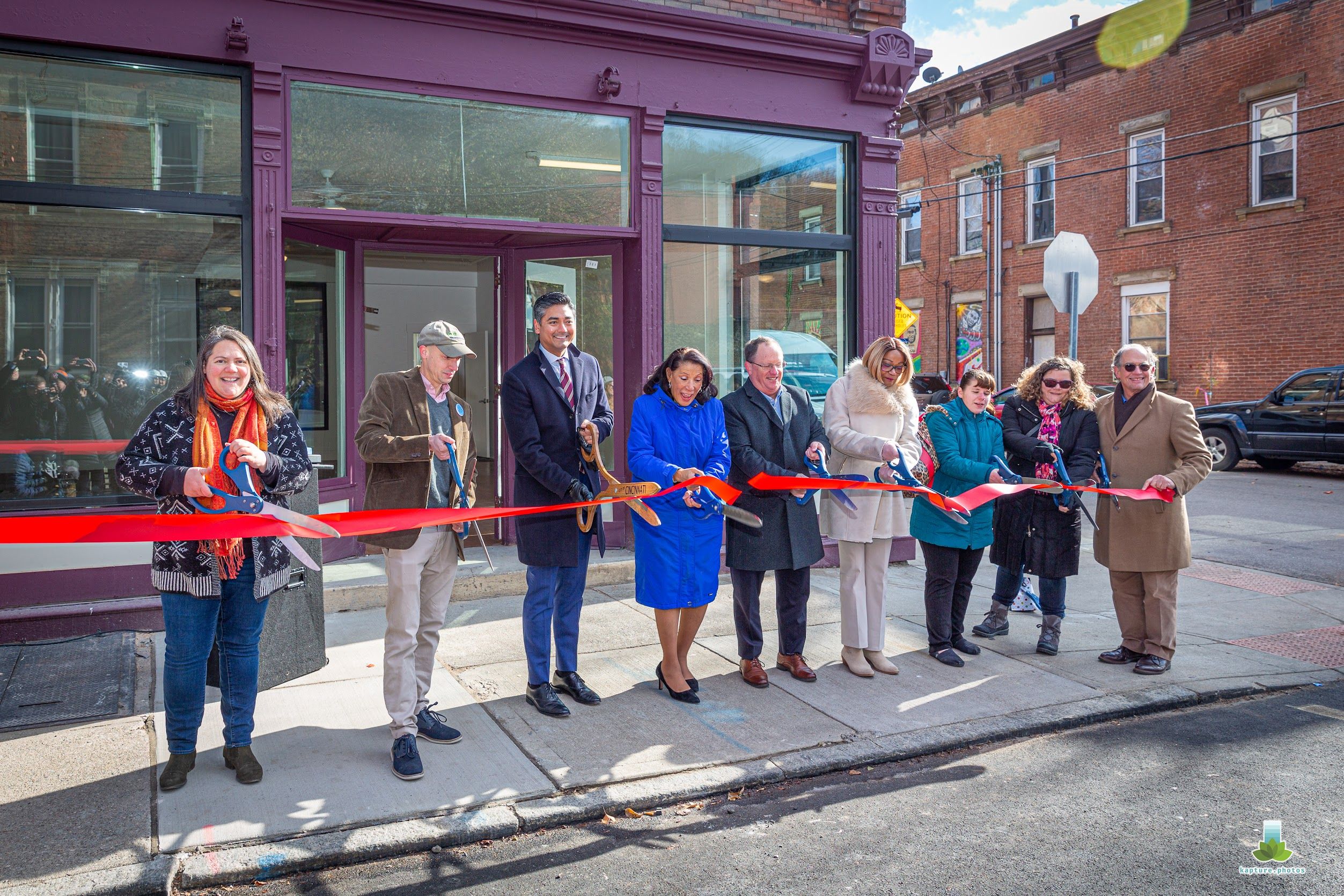 Ribbon-cutting ceremony outside a purple storefront. A diverse group of people in coats and scarves holds red ribbon and blue scissors, with brick buildings in the background.
