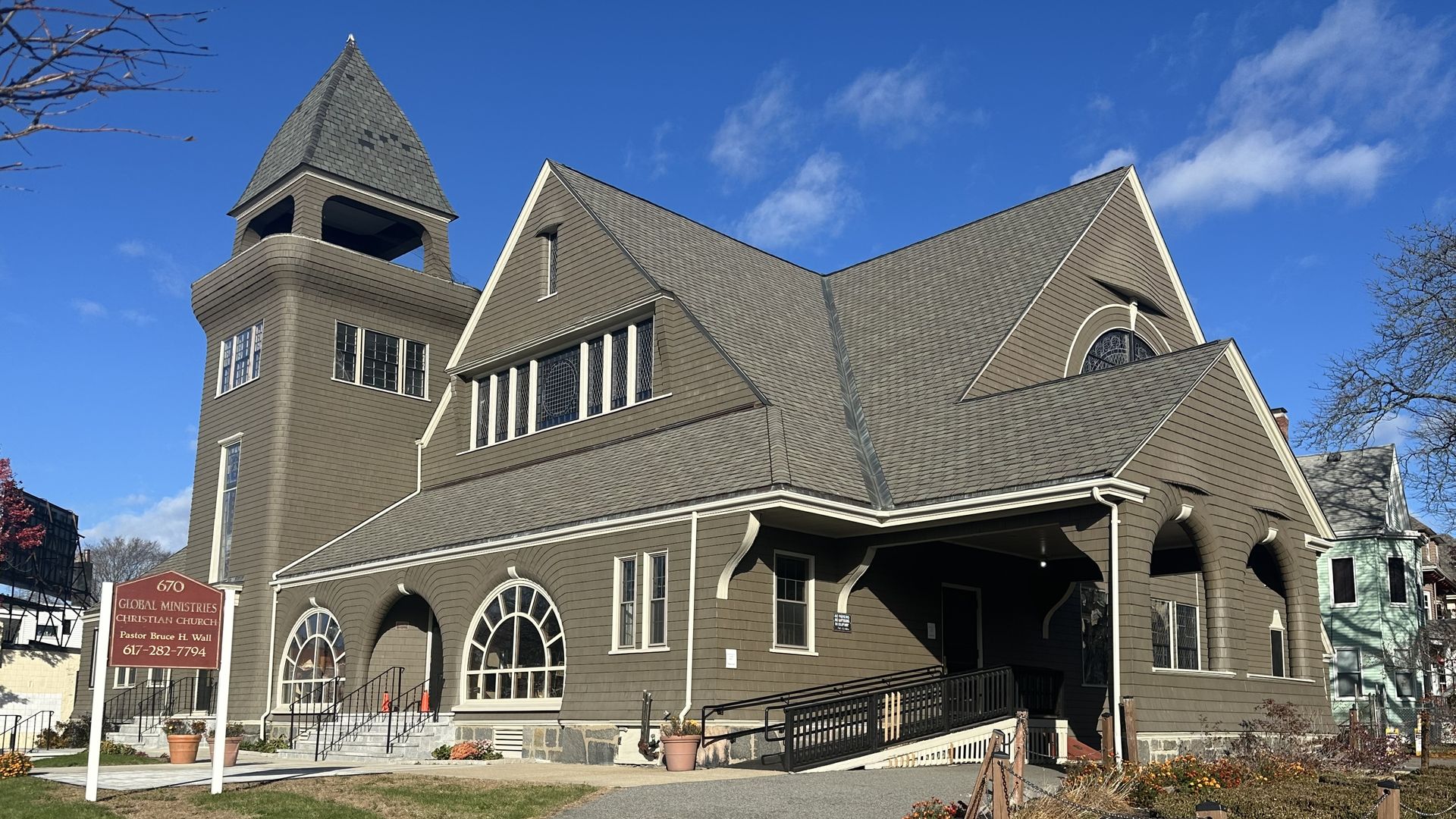 Brown church building with steep roof, arched windows, and a tower, under a blue sky. Sign reads Global Ministries Christian Church, Pastor Bruce H. Wall, with phone number.
