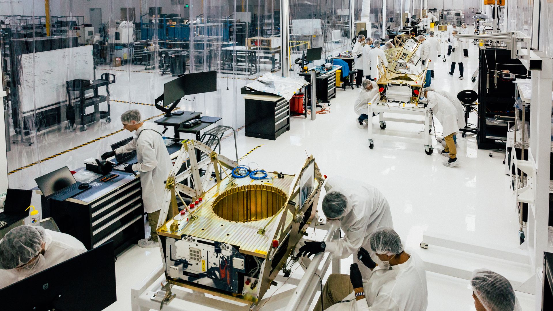 Technicians in white lab coats and hairnets work on gold-colored satellite components in a clean, white, high-tech laboratory with transparent partitions and multiple workstations.