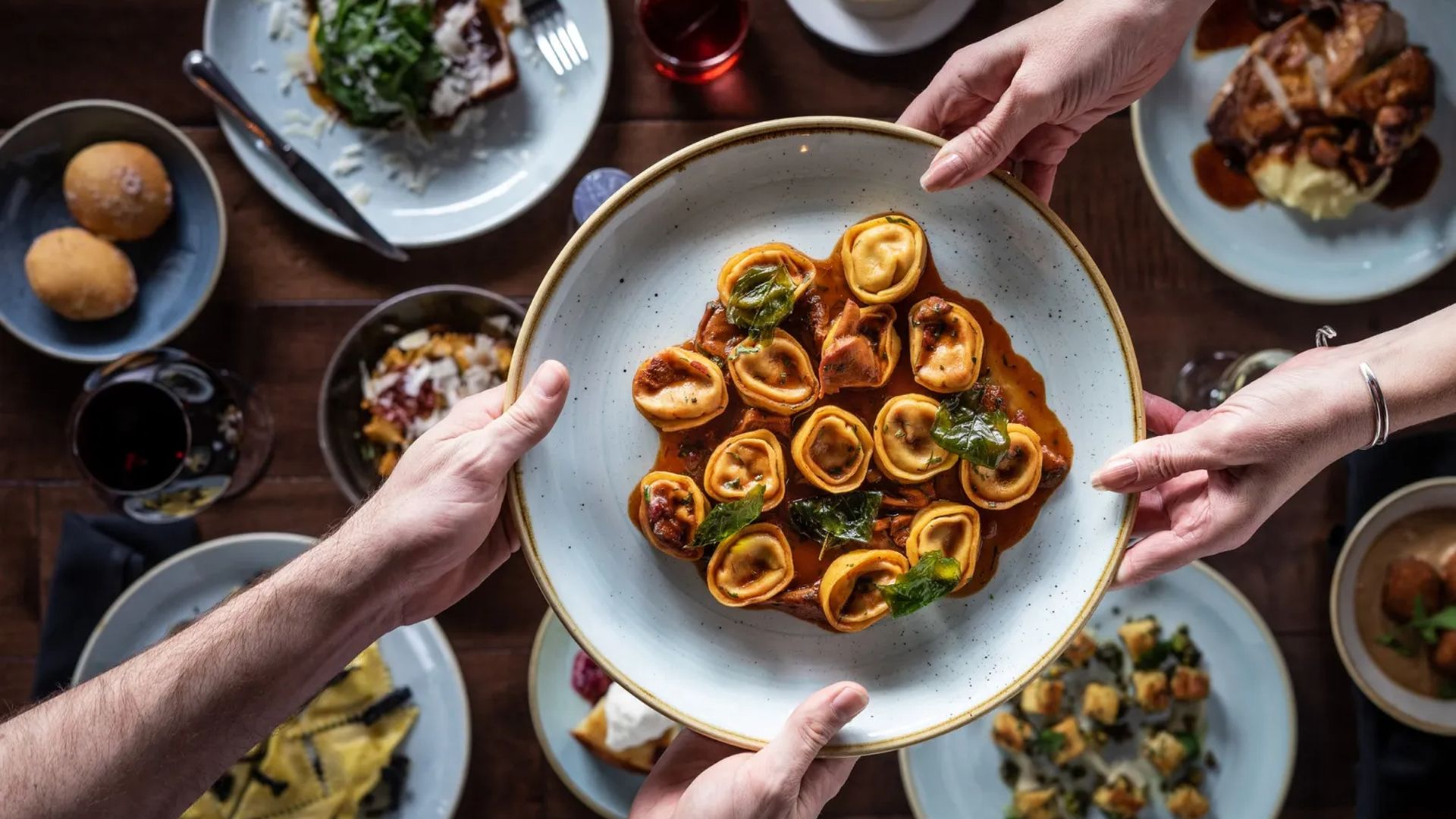Top-down view of a group sharing a large bowl of tortellini in tomato sauce, garnished with greens, held by several hands over a wooden table with plates of bread, salad, and other dishes.