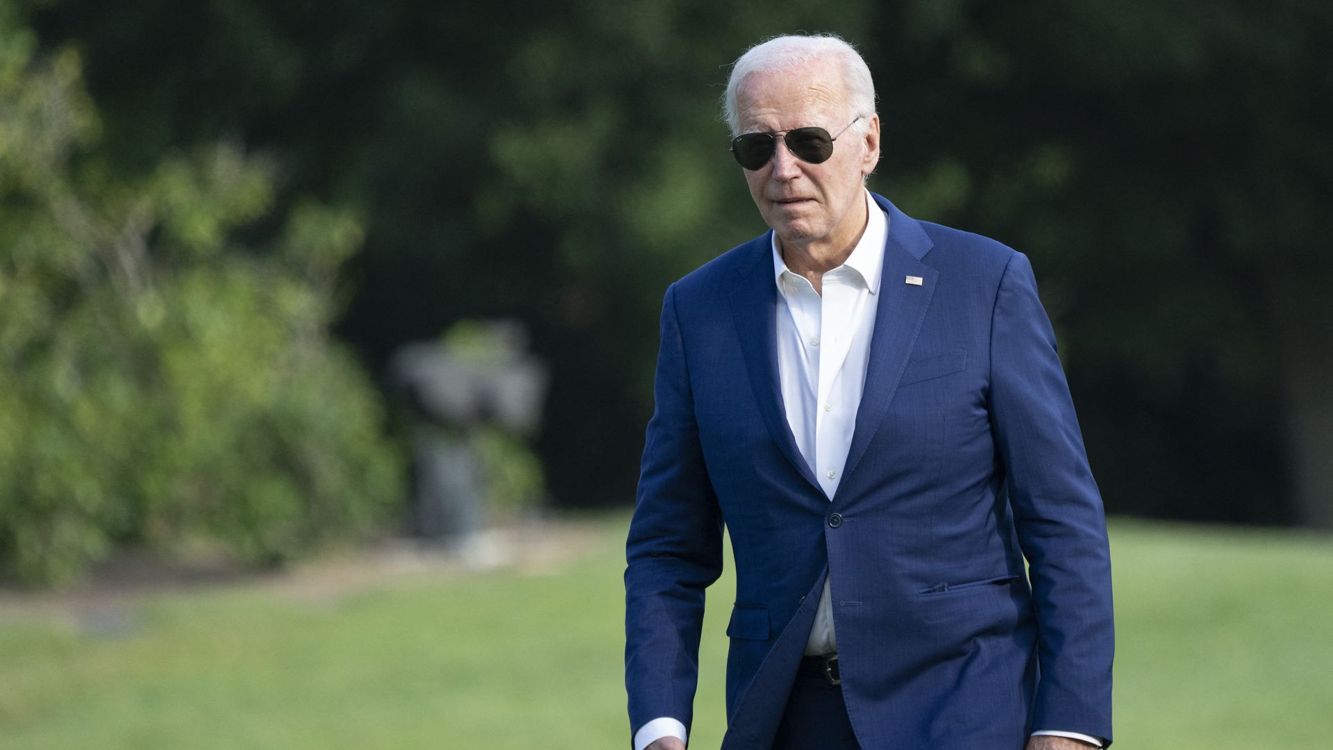 Biden walks down a lawn. He wears a blue suit and white shirt and aviator sunglasses. 