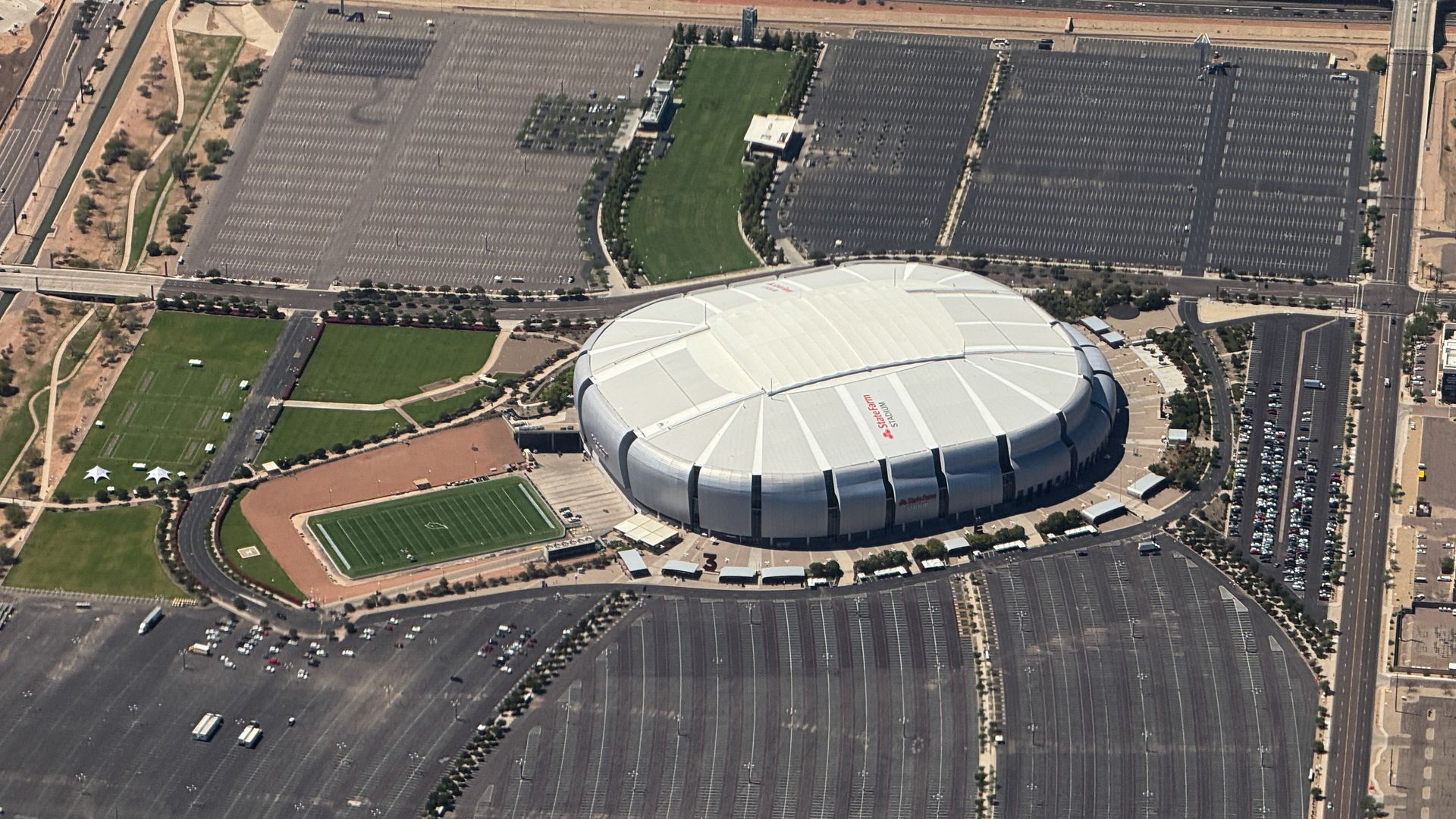 An aerial image from above a light silver, oblong football stadium surrounded by mostly empty parking lots. 