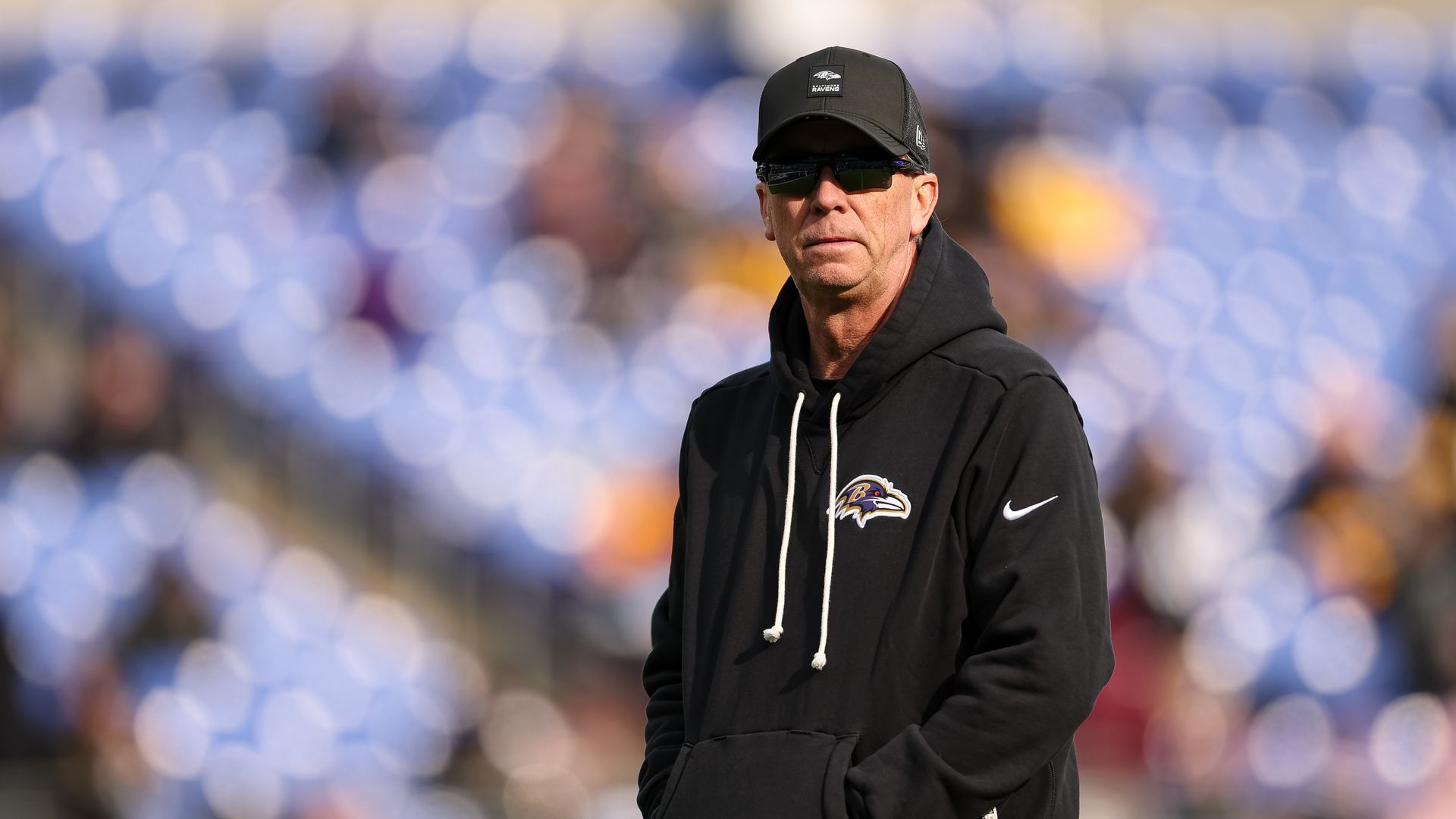 Man wearing black Baltimore Ravens hoodie and cap with sunglasses, standing outdoors with blurred blue stadium seats and crowd background.