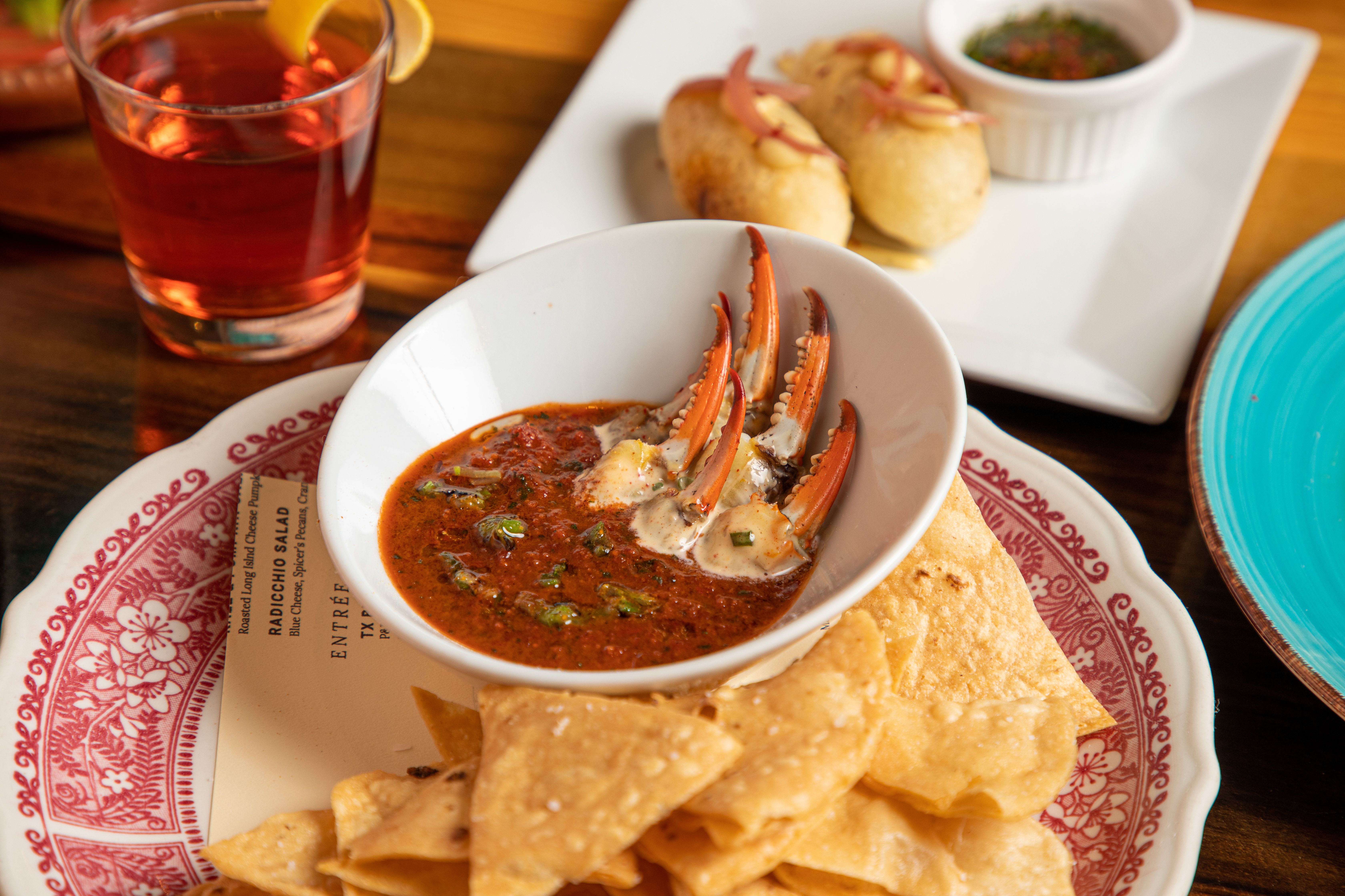 A small white bowl filled with a red sauce and crab claws sits atop a red-rimmed plate with tortilla chips.