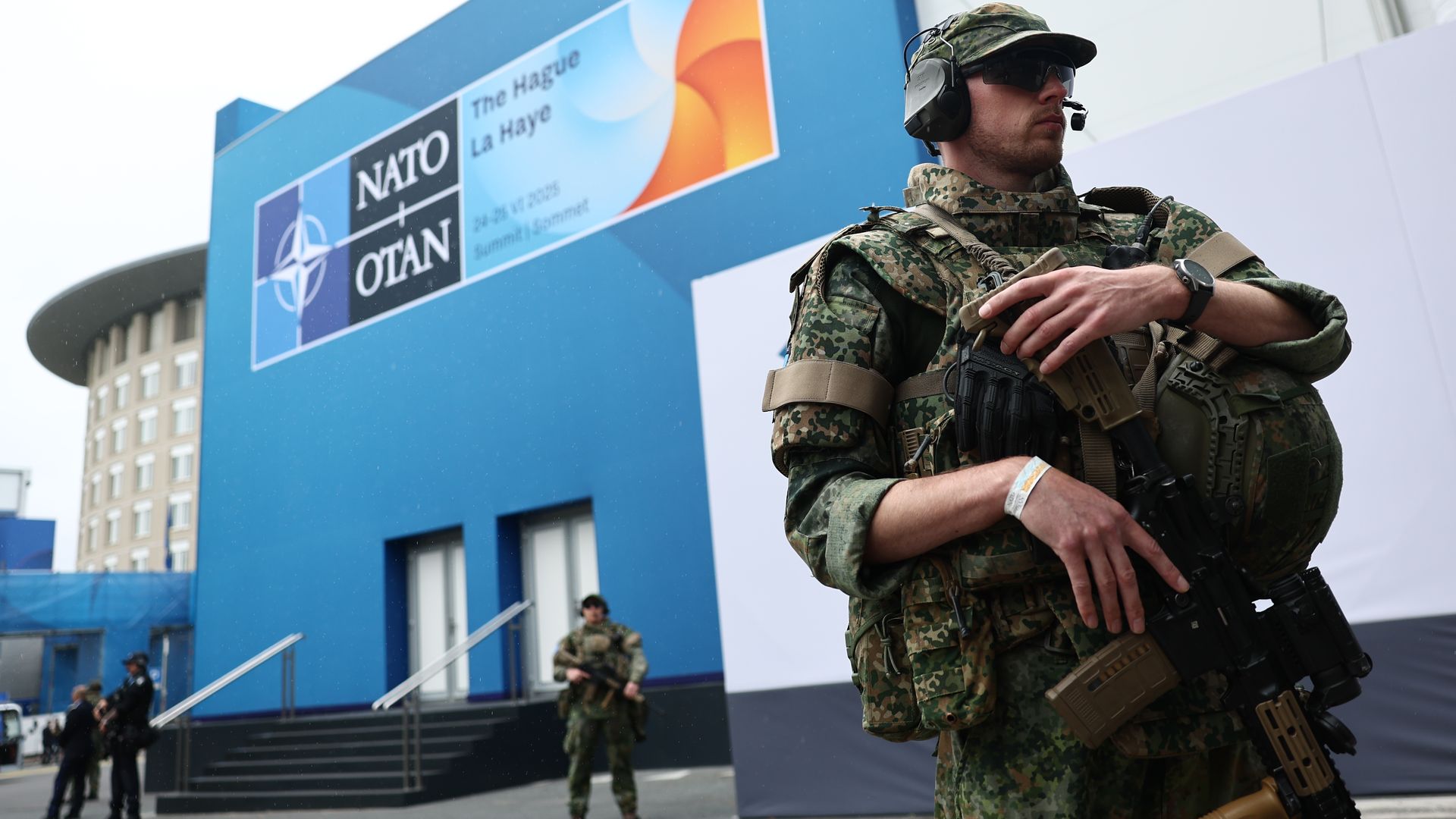 Armed military guards stand outside the NATO summit building in the Netherlands. The building is blue with the NATO-OTAN logo on the side.