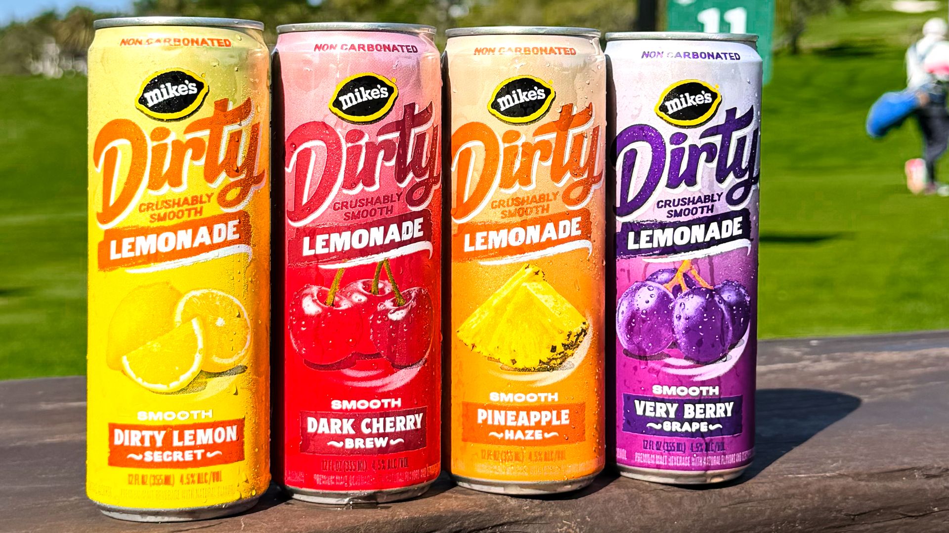Four colorful cans of Mike's Dirty Lemonade arranged in a row on a wooden table outdoors; yellow lemon, red cherry, orange pineapple, and purple grape flavors, with a park setting in the background.