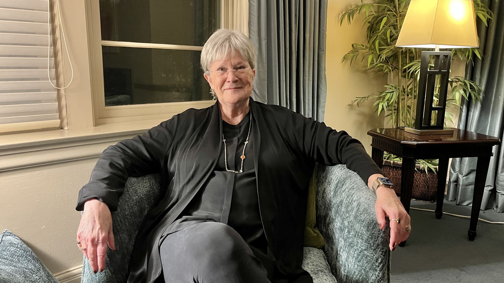 Vivien Jennings of Rainy Day Books sits with short gray hair and glasses on a gray armchair wearing black clothes and silver jewelry, smiling in a back room of Unity Temple on the Plaza.