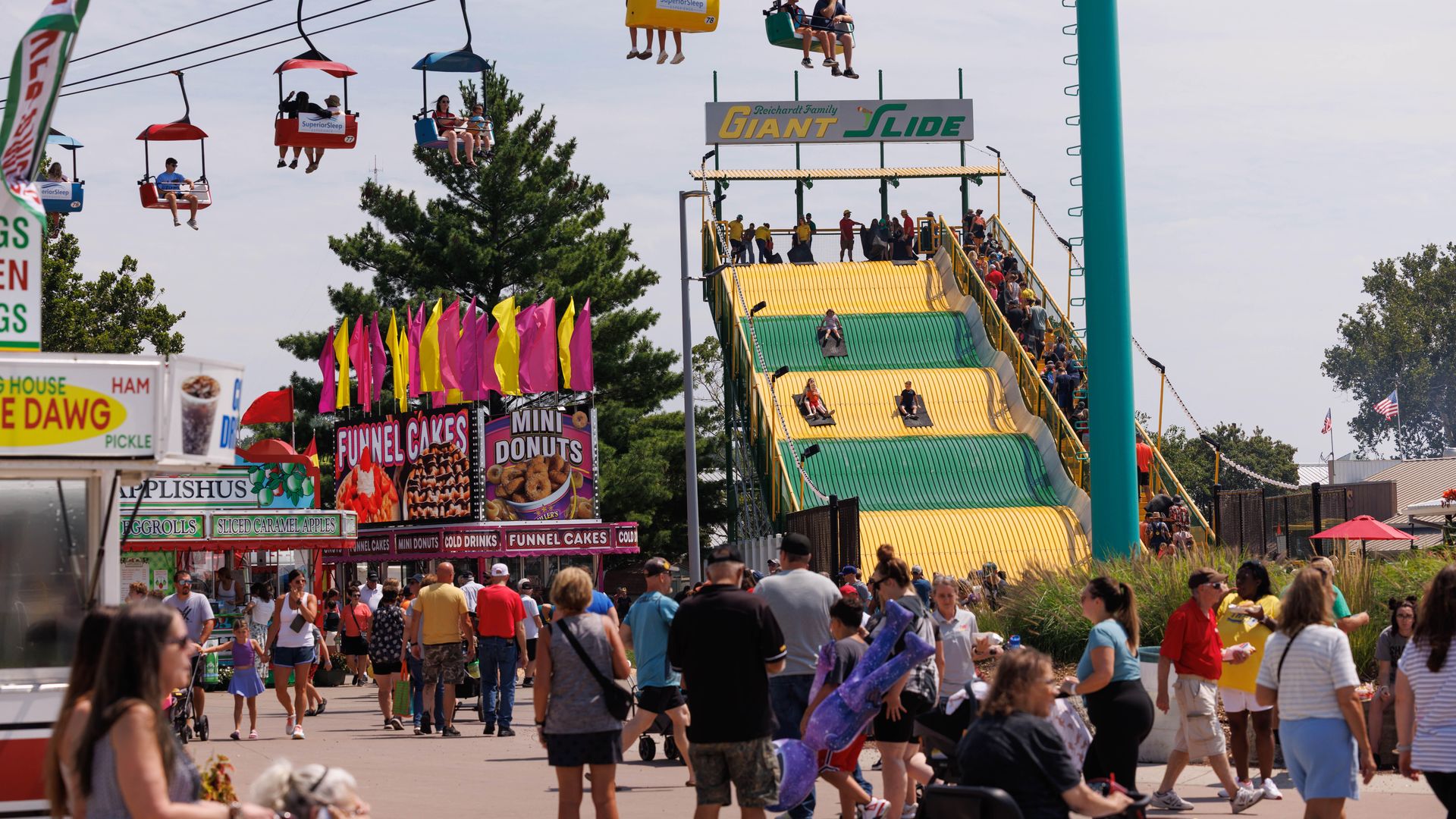 Crowded carnival scene with a towering slide ride in yellow and green, a colorful chairlift of riders overhead, and a large "GIANT SLIDE" banner. People walk past food stalls on a sunny day.
