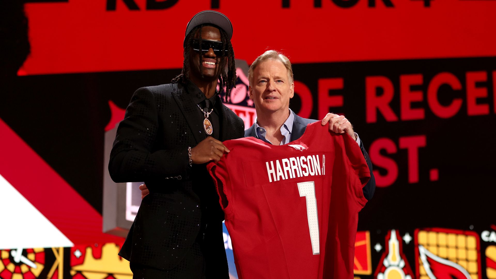 Two men hold up a red football jersey with the name Harrison on the back. 