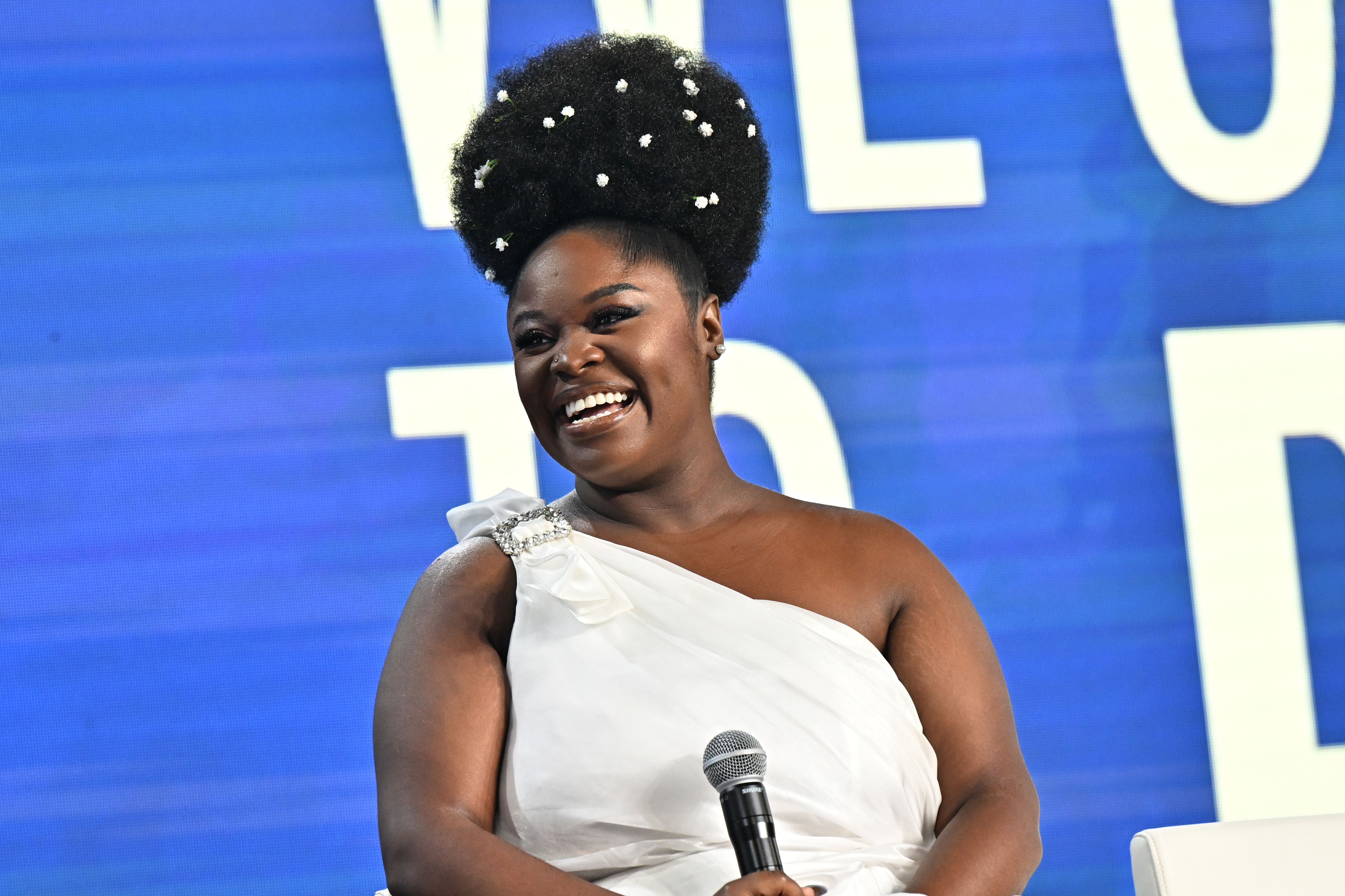 Smiling woman in a white one-shoulder dress with flower-decorated afro hairstyle holding a microphone, sitting against a blue background with large white text.