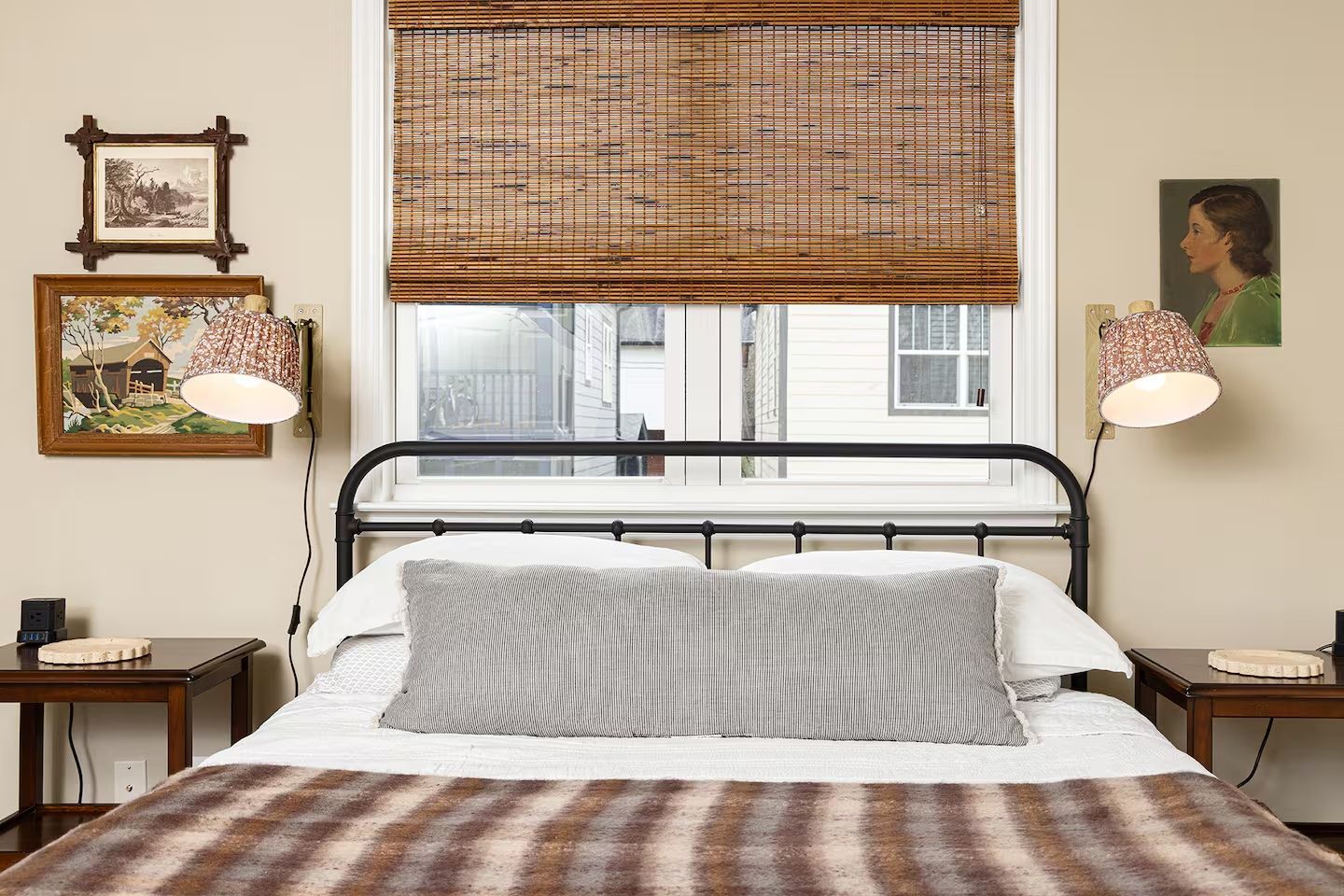 Bedroom with a black metal bed frame, white bedding, long gray pillow, brown plaid blanket, bamboo window shade, two wooden nightstands, floral lamps, and framed art on beige walls.