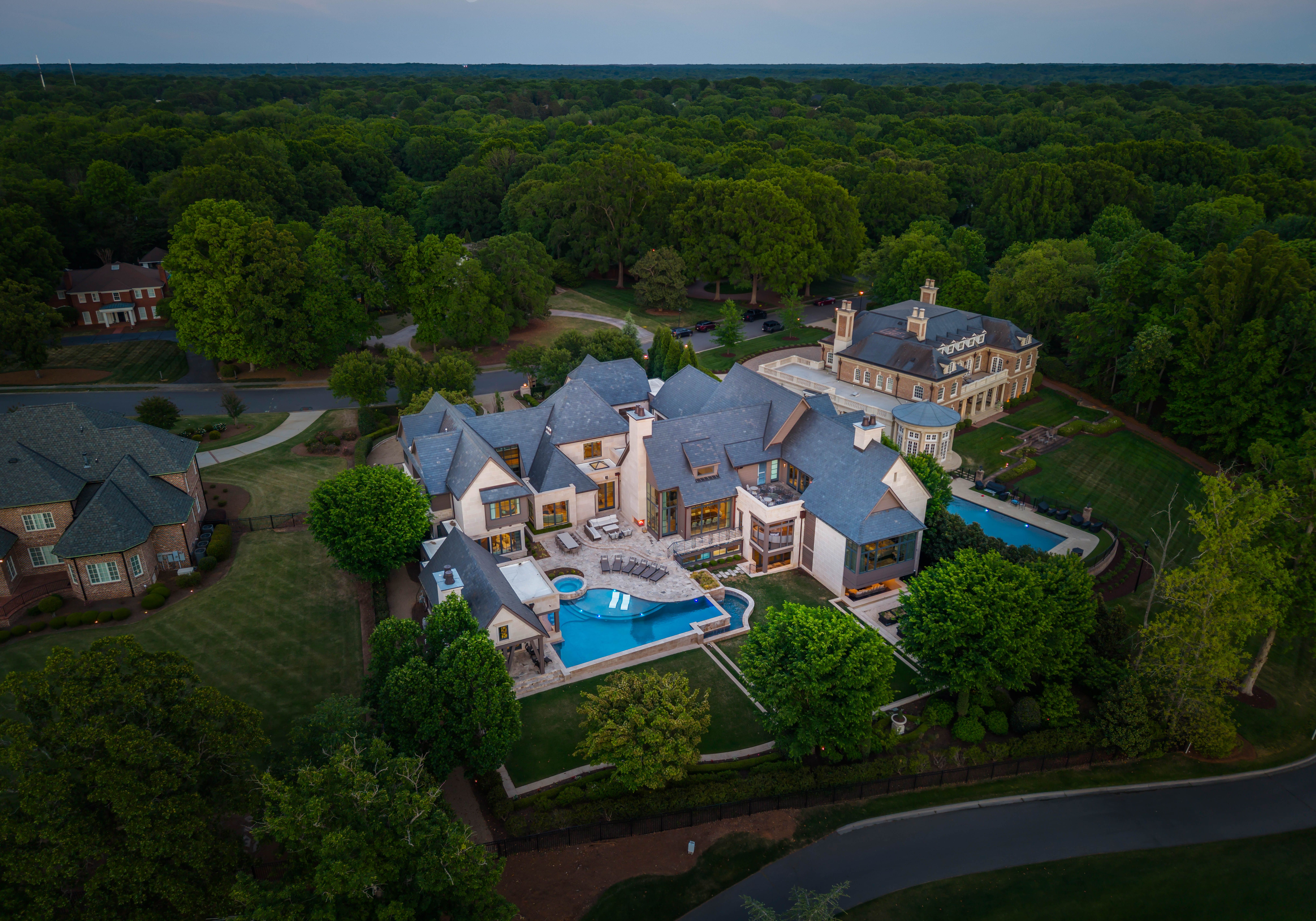 Aerial view of a large mansion complex with gray slate roofs, beige stone facades, and a bright blue pool. Surrounded by dense green forest and manicured lawns; curved road at the edge.