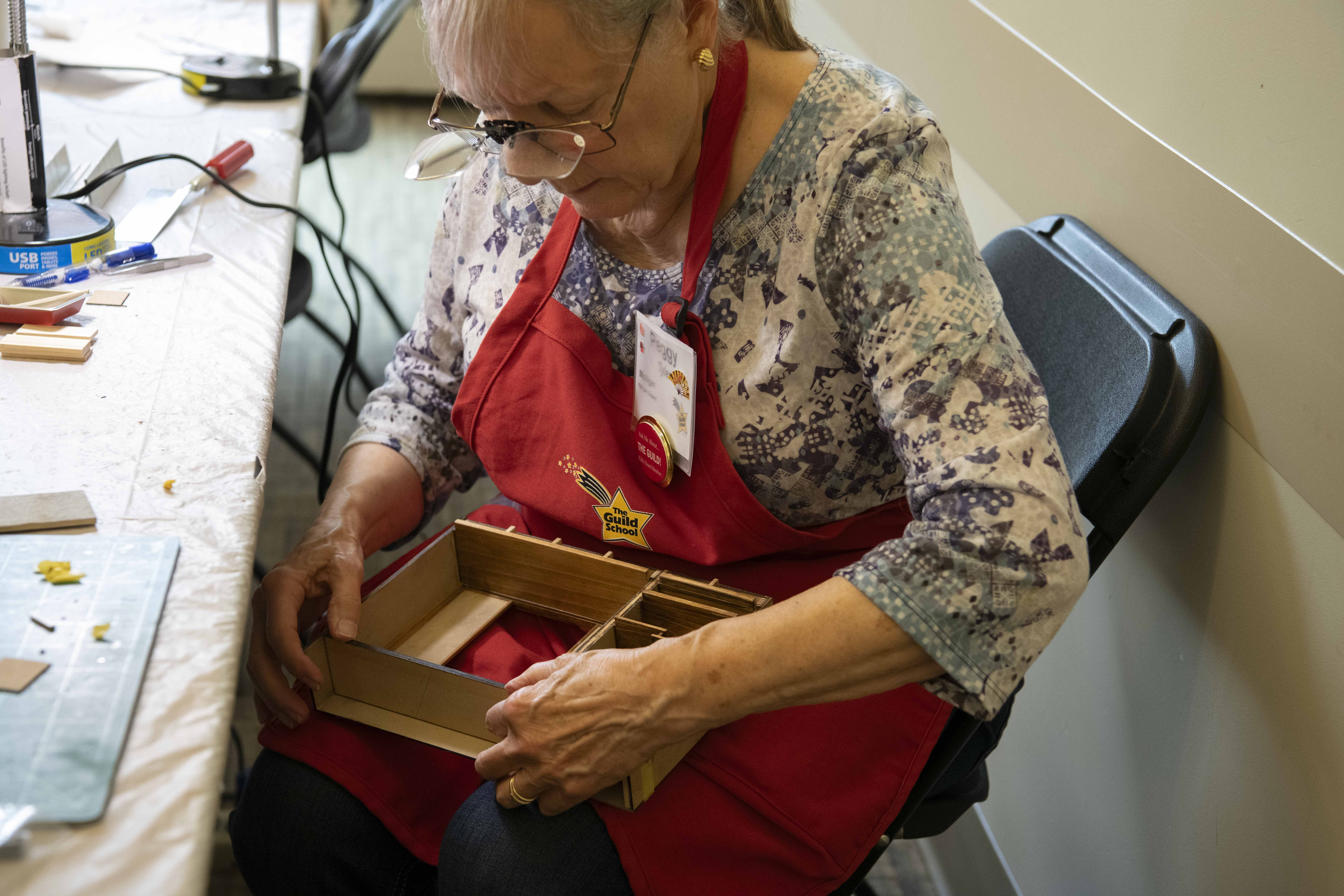 An elderly woman in a red apron and patterned shirt sits at a workshop table, looking down at wooden pieces in a box, wearing glasses and a badge, with craft tools and cords nearby.