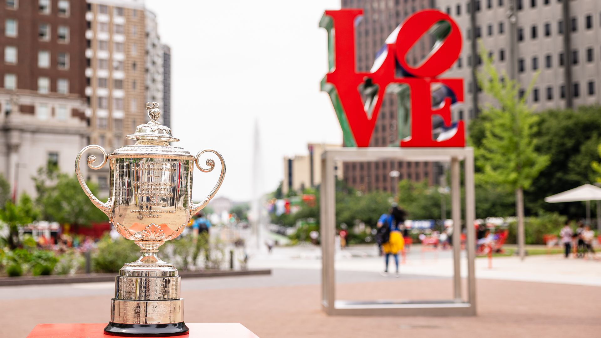 Shiny silver trophy on a pedestal in a city square, with a large red "LOVE" sculpture and blurred buildings in the background; a bird flies overhead and pedestrians stroll.