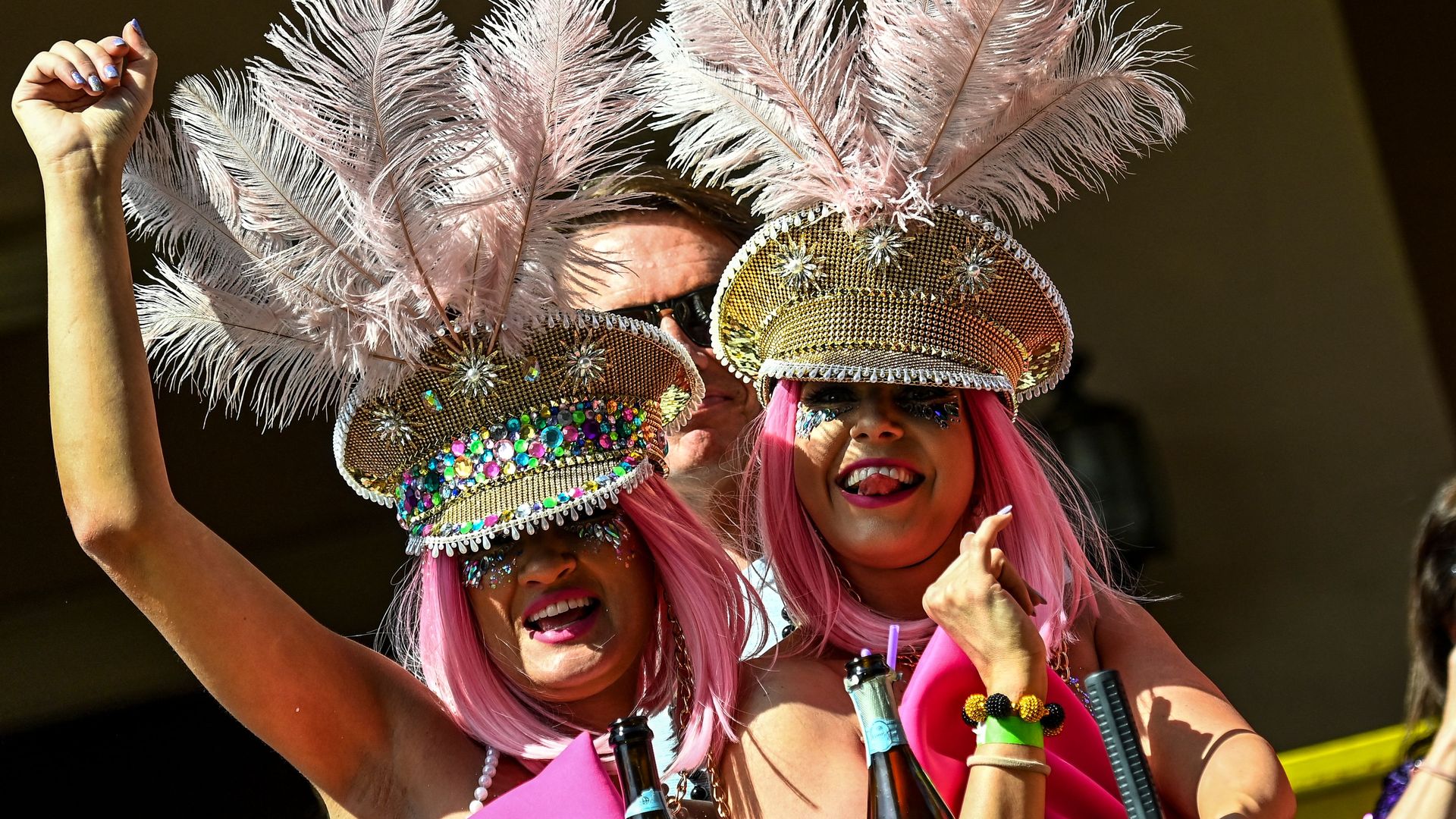 Two women, wearing festooned, glittery hats and pink wigs, smile from a Zulu parade float.