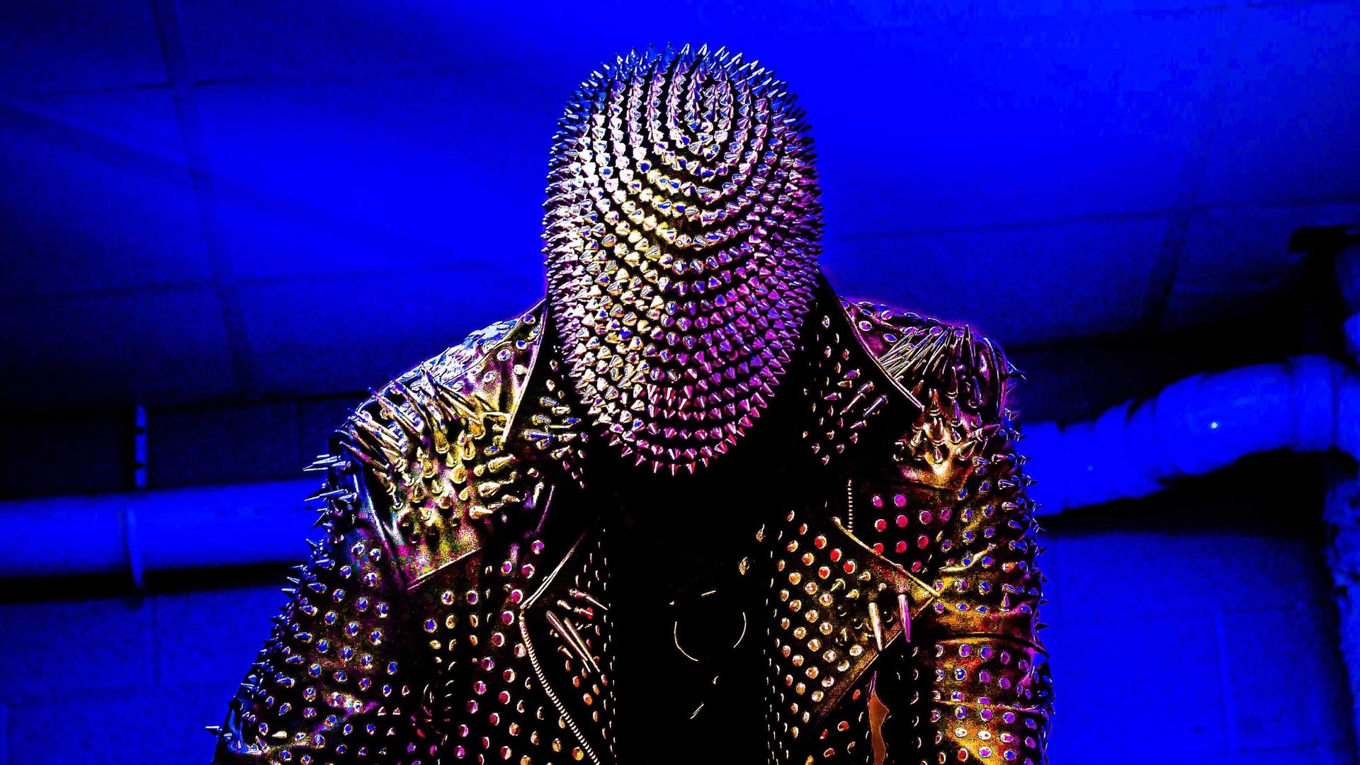 A man in a spiked mask and leather jacked in front of neon background.