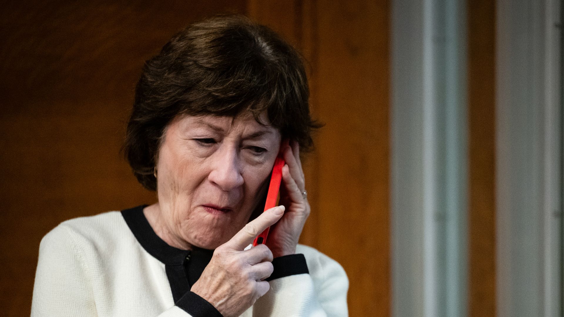 Sen. Collins, with pursed lips, cradles a cell phone in a red protective case with both hands